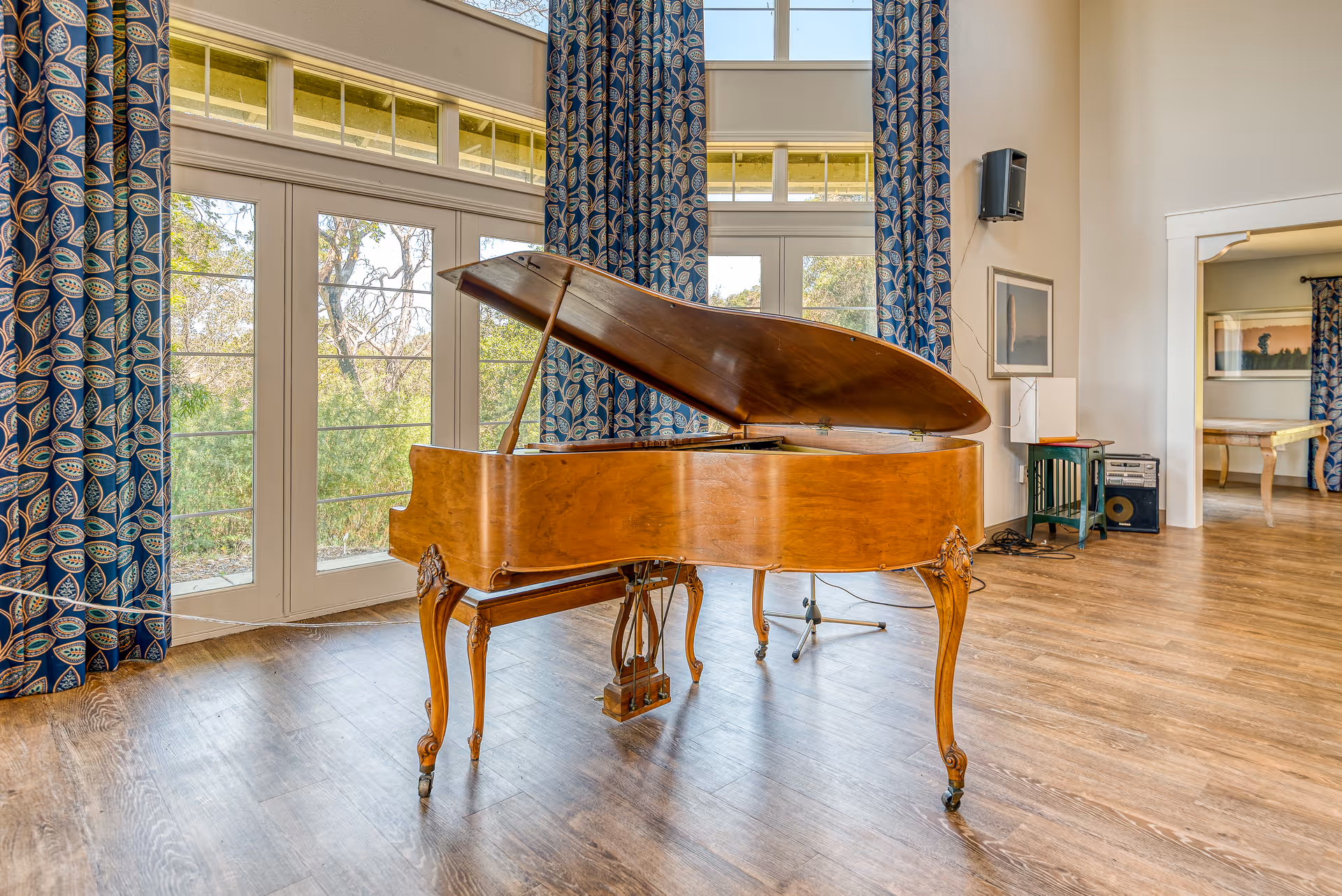 A wooden grand piano with its lid open sits on a polished wooden floor in a bright room with large windows and blue patterned curtains. The room has high ceilings and light-colored walls, with a small table and speaker visible in the background.