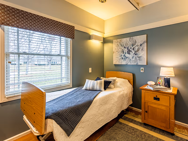 A cozy bedroom with a single bed featuring a wooden headboard and footboard, white quilted bedding, and several pillows including a striped one. Next to the bed is a wooden nightstand with a lamp, books, and decorative items. A large window with blinds and a patterned valance lets in natural light. The walls are painted a muted blue-gray, and a large floral artwork hangs above the bed. A striped rug covers the floor.