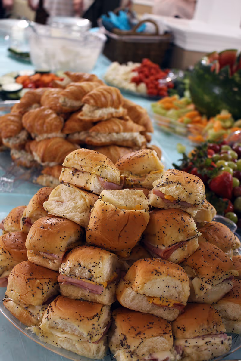 A close-up view of a large platter filled with small ham and cheese sandwiches on poppy seed buns, with a background showing a pile of croissant sandwiches and various trays of fresh cut vegetables and fruit.