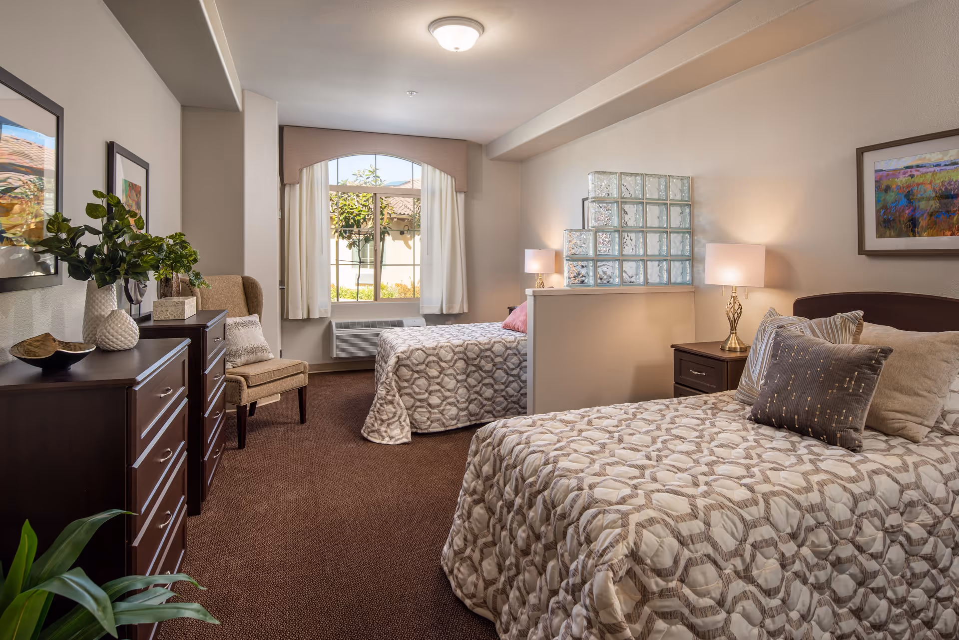 A well-lit bedroom with two beds separated by a half wall with glass blocks. Each bed has patterned bedding and pillows. There are two nightstands with lamps, a window with curtains letting in natural light, a chair, two dressers with decorative items, and framed artwork on the walls.