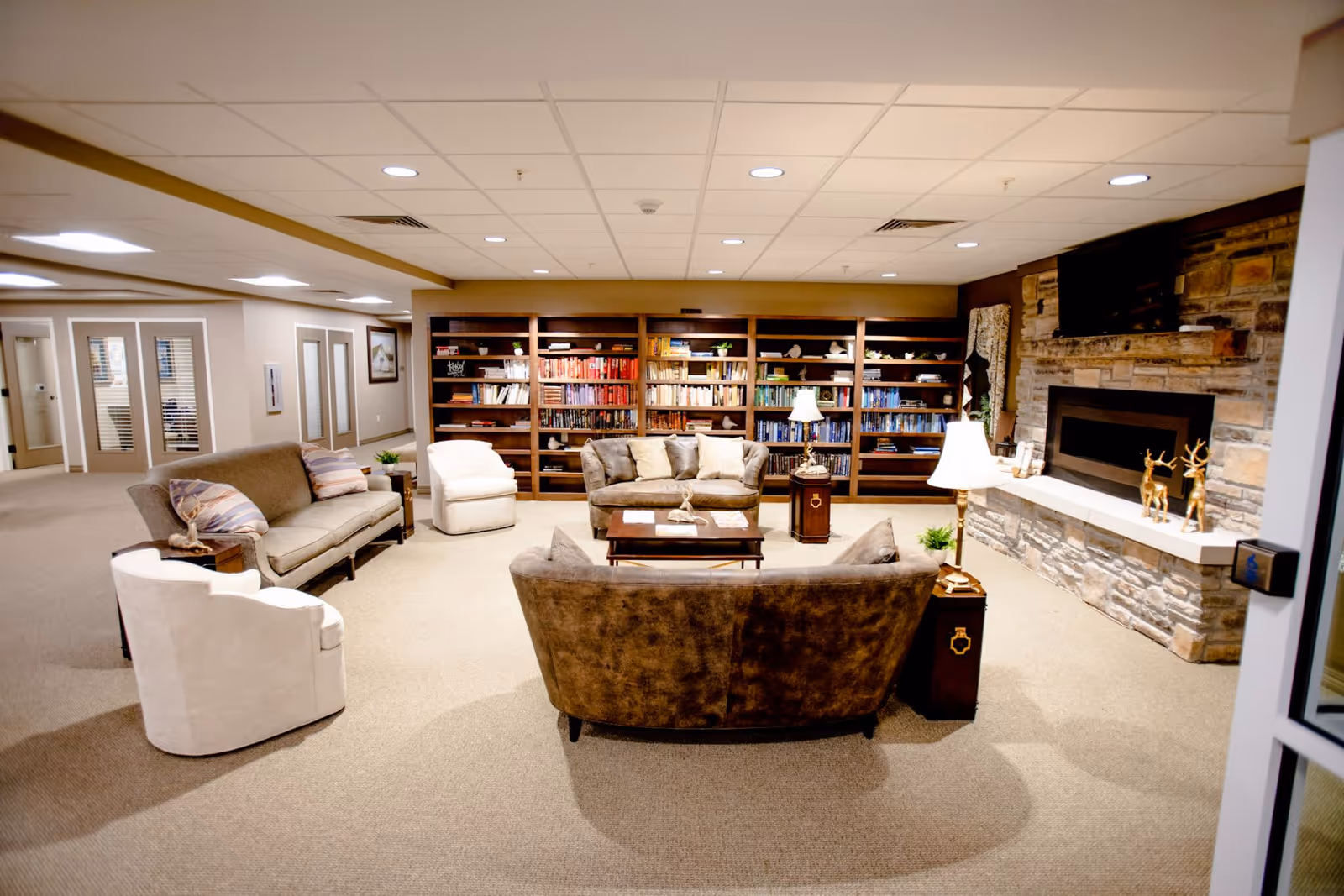 A cozy living room area with multiple sofas and armchairs arranged around a coffee table. Behind the seating is a large bookshelf filled with books and decorative items. To the right, there is a stone fireplace with a mounted TV above it and decorative golden deer figurines on the mantel. The room has a beige carpet and a drop ceiling with recessed lighting.