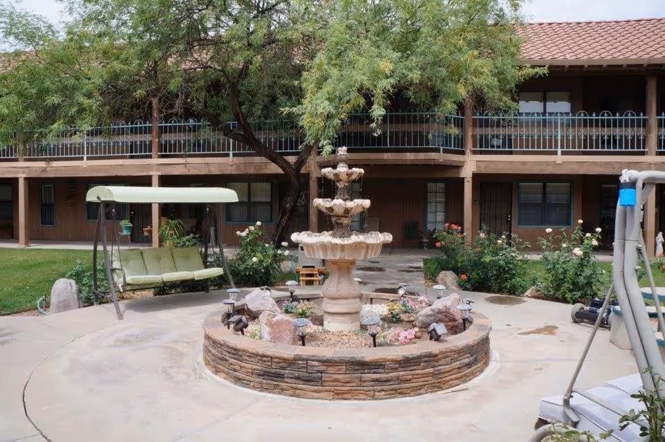 Outdoor courtyard area with a multi-tiered stone fountain surrounded by flowers and rocks. There is a green cushioned swing on the left and a building with a tiled roof and balcony in the background. Trees and bushes are also visible around the courtyard.