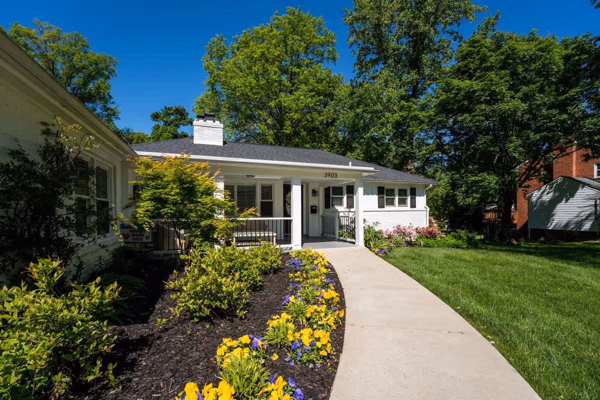 White, single-story senior living building with a front porch, curved walkway, and landscaped garden.