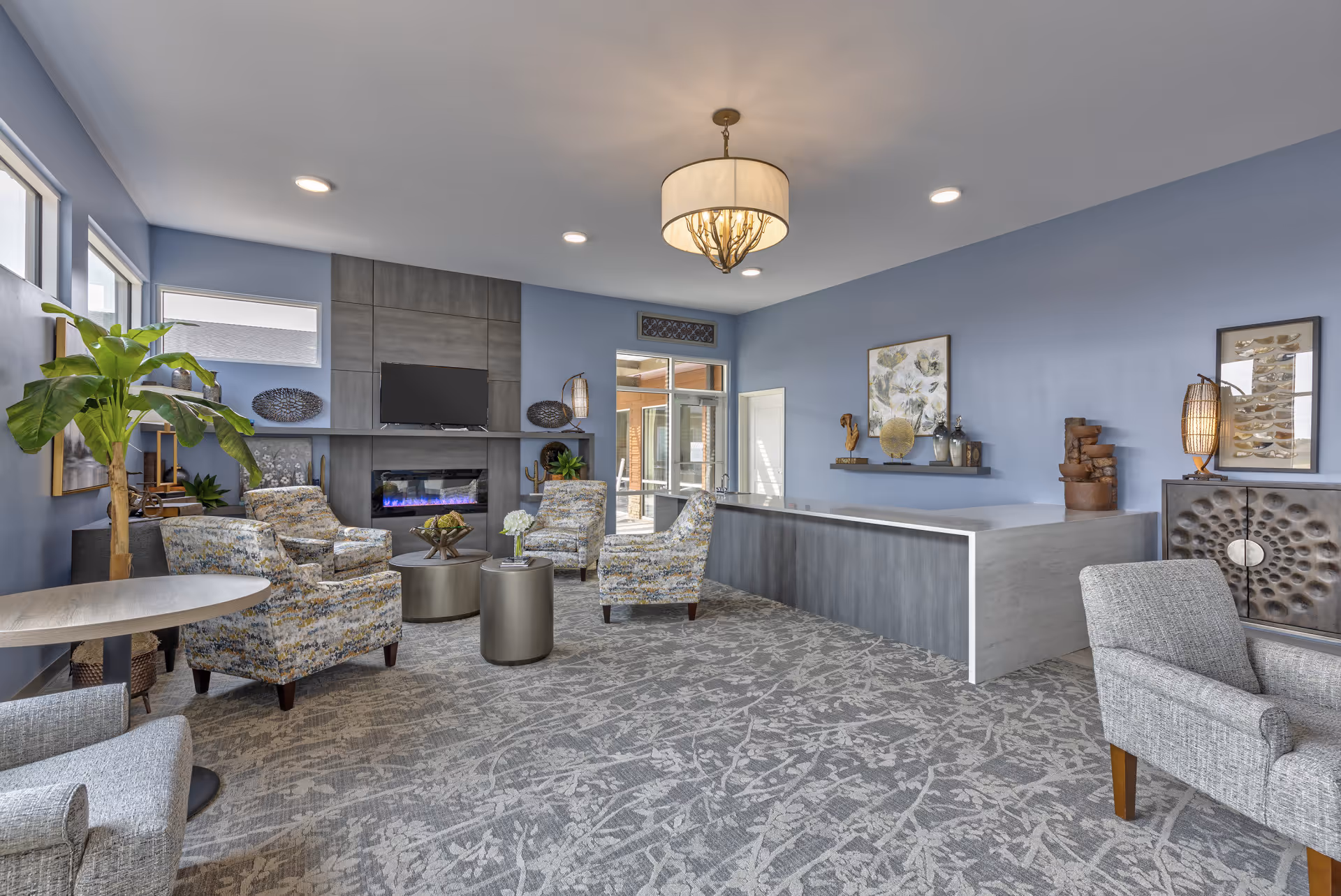 A modern living room area with light blue walls and patterned carpet. The room features several upholstered armchairs arranged around two round tables in front of a fireplace with a mounted TV above it. There are decorative plants, artwork, and sculptures on shelves and furniture. A large light fixture hangs from the ceiling, and windows allow natural light into the space.