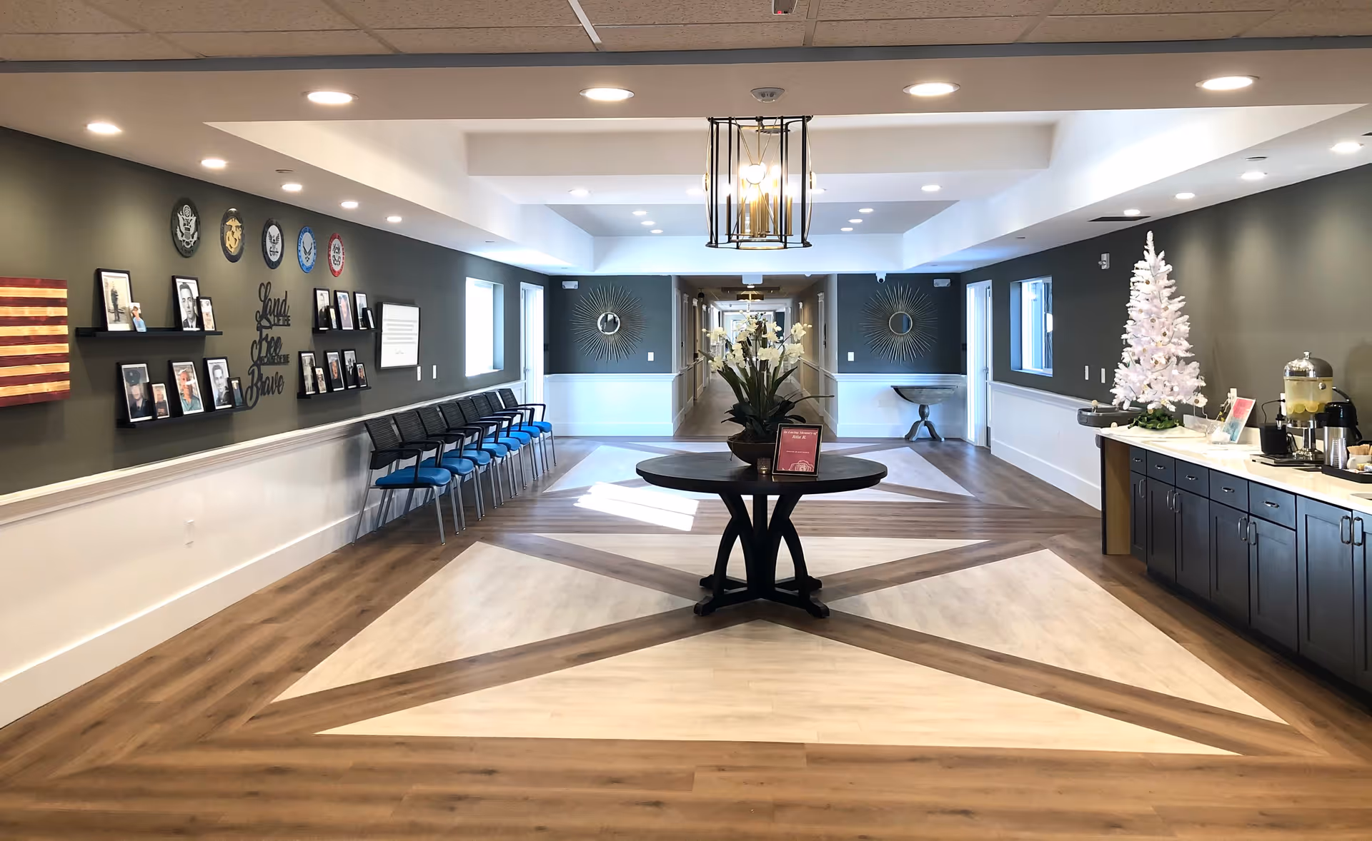 A spacious hallway in a senior living facility with a decorative star pattern on the wooden floor. A round table with a flower arrangement is centered in the foreground. On the left wall, there are framed photos, military emblems, and a sign that reads 'Land of the Free Brave' above a row of blue and black chairs. The right side features a countertop with a white Christmas tree, a beverage dispenser, and other refreshments. The hallway extends into the distance with doors and decorative mirrors on the walls.