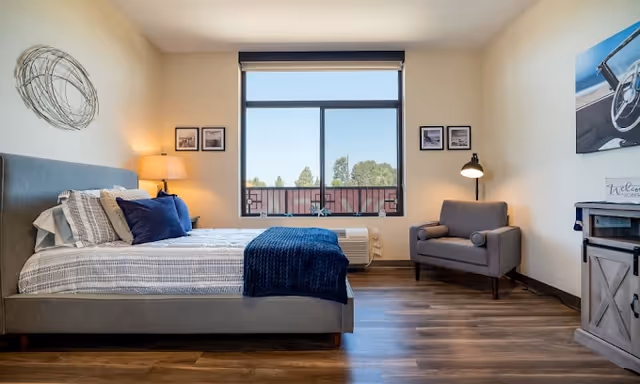 A bright and modern bedroom with a large window showing trees outside. The room features a neatly made bed with blue and gray bedding, a gray upholstered armchair with a floor lamp beside it, wooden flooring, and framed pictures on the walls.