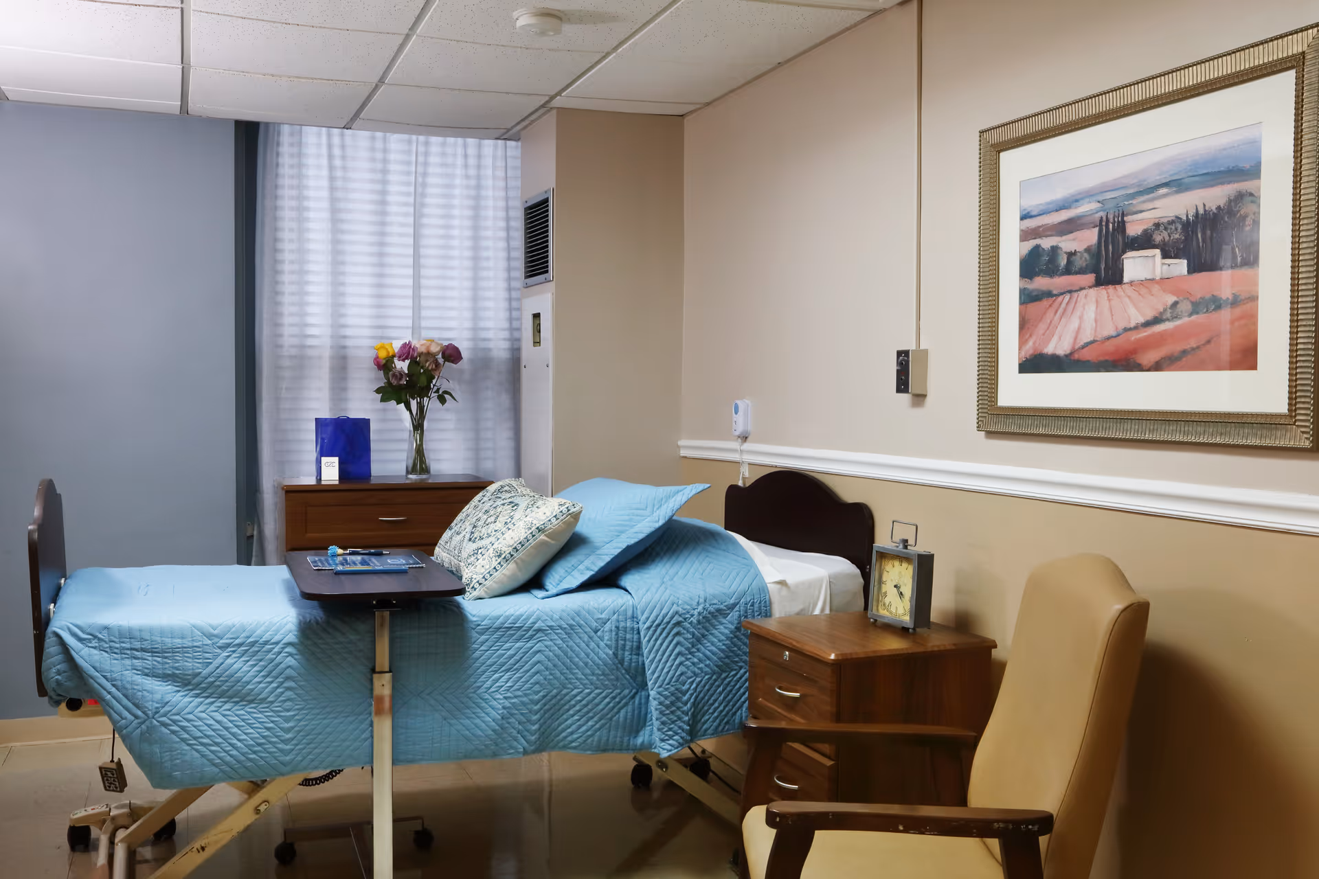 A hospital-style bed with blue bedding and pillows in a room with beige walls. Next to the bed is a wooden nightstand with a clock on it, and a beige armchair is positioned nearby. A framed landscape painting hangs on the wall above the nightstand. In the background, there is a window with white curtains and a small wooden dresser with a vase of flowers and a blue gift bag on top.