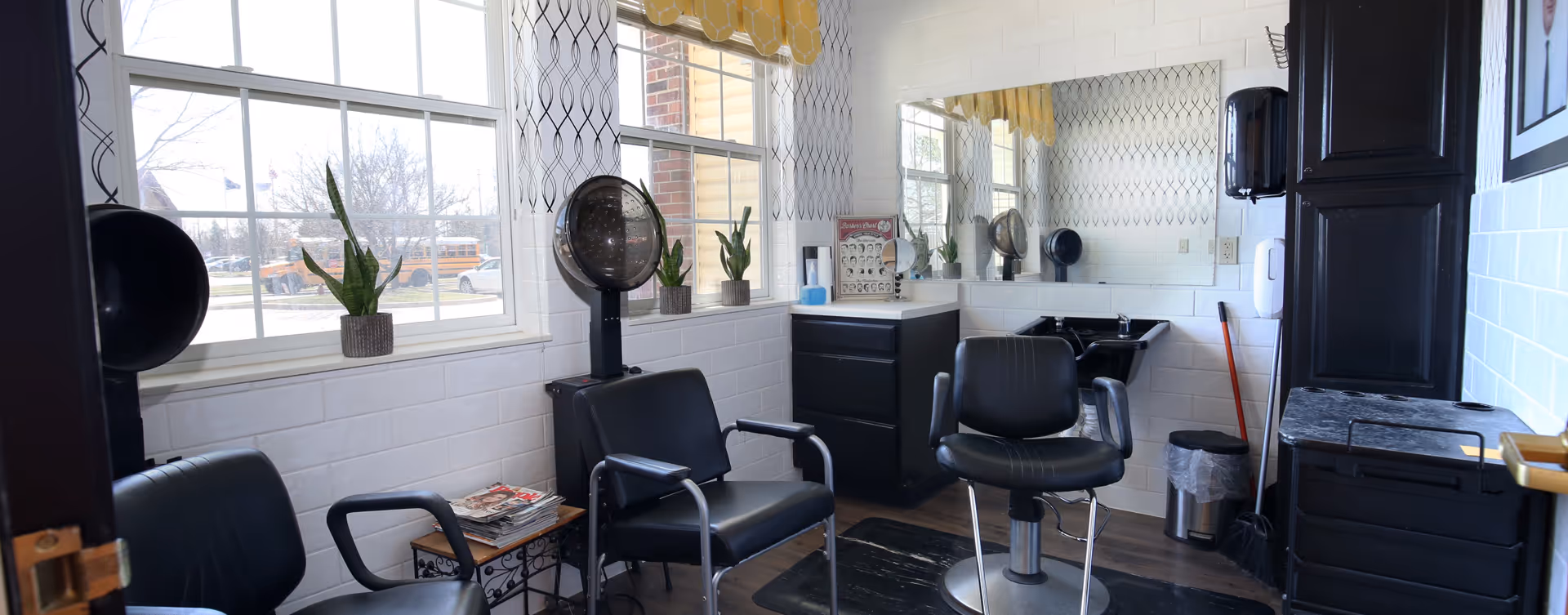 Interior of a small salon or hair styling room with black salon chairs, a hair dryer, a large mirror on the wall, a sink for washing hair, and potted plants on the windowsill. The room has white tiled walls and large windows letting in natural light.