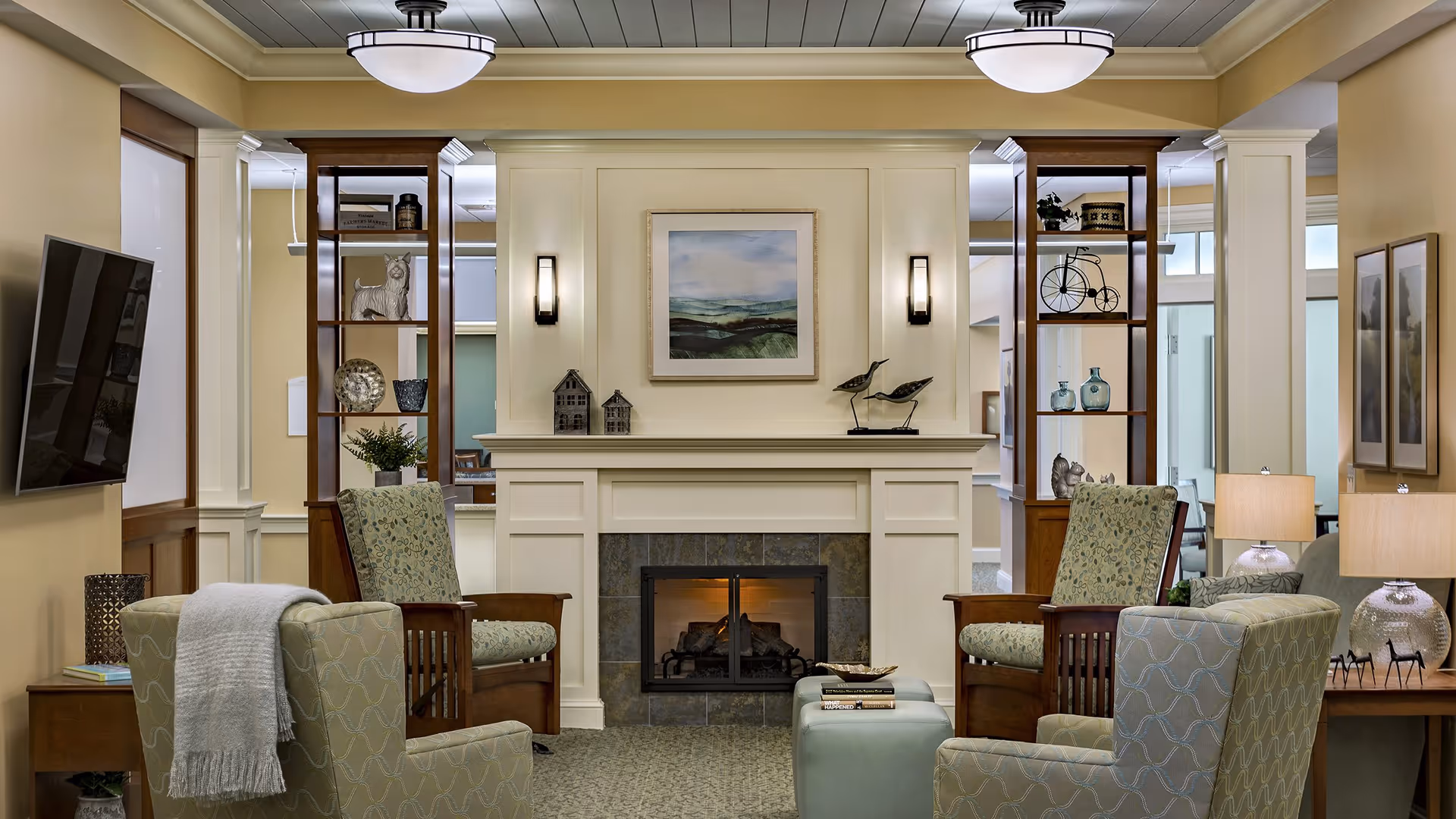 A cozy living room area with a central fireplace surrounded by four upholstered armchairs. Two wooden shelving units flank the fireplace, decorated with various ornaments and plants. A flat-screen TV is mounted on the left wall, and two table lamps with beige shades are on side tables. The walls are painted a warm beige color, and a framed landscape painting hangs above the fireplace.