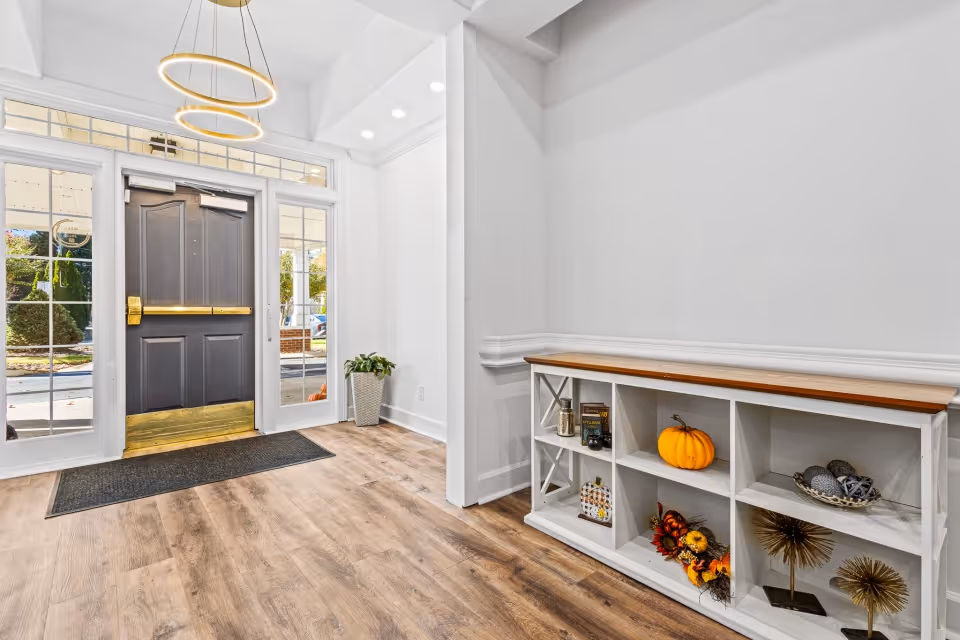 Bright entrance lobby with an open black front door, wood-look flooring, modern ring pendant lights, and a white cubby shelf decorated with seasonal items including a pumpkin.