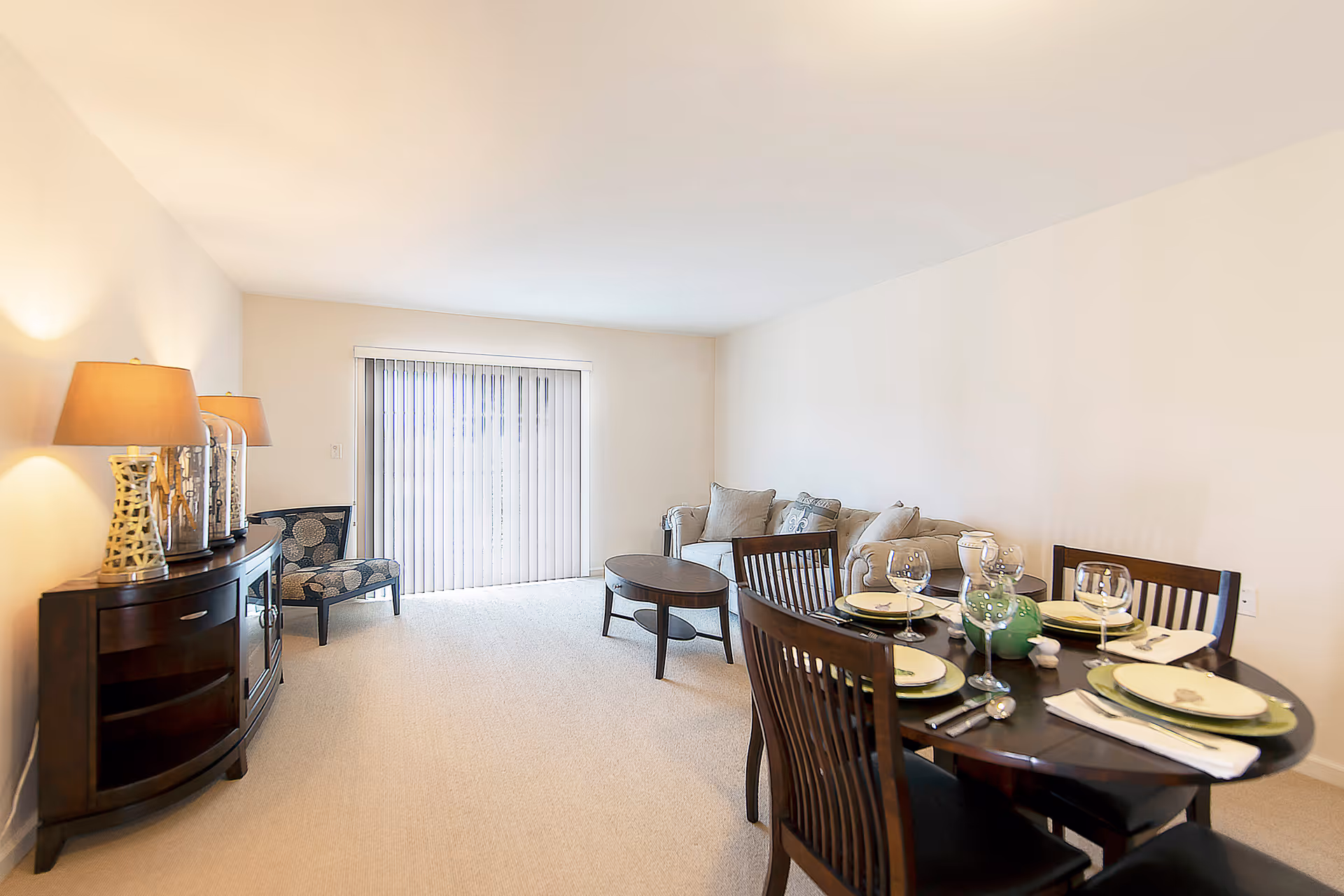 Open-plan living and dining room with a sofa and coffee table, a dining table set for four, and a sideboard with lamps in front of sliding glass doors.