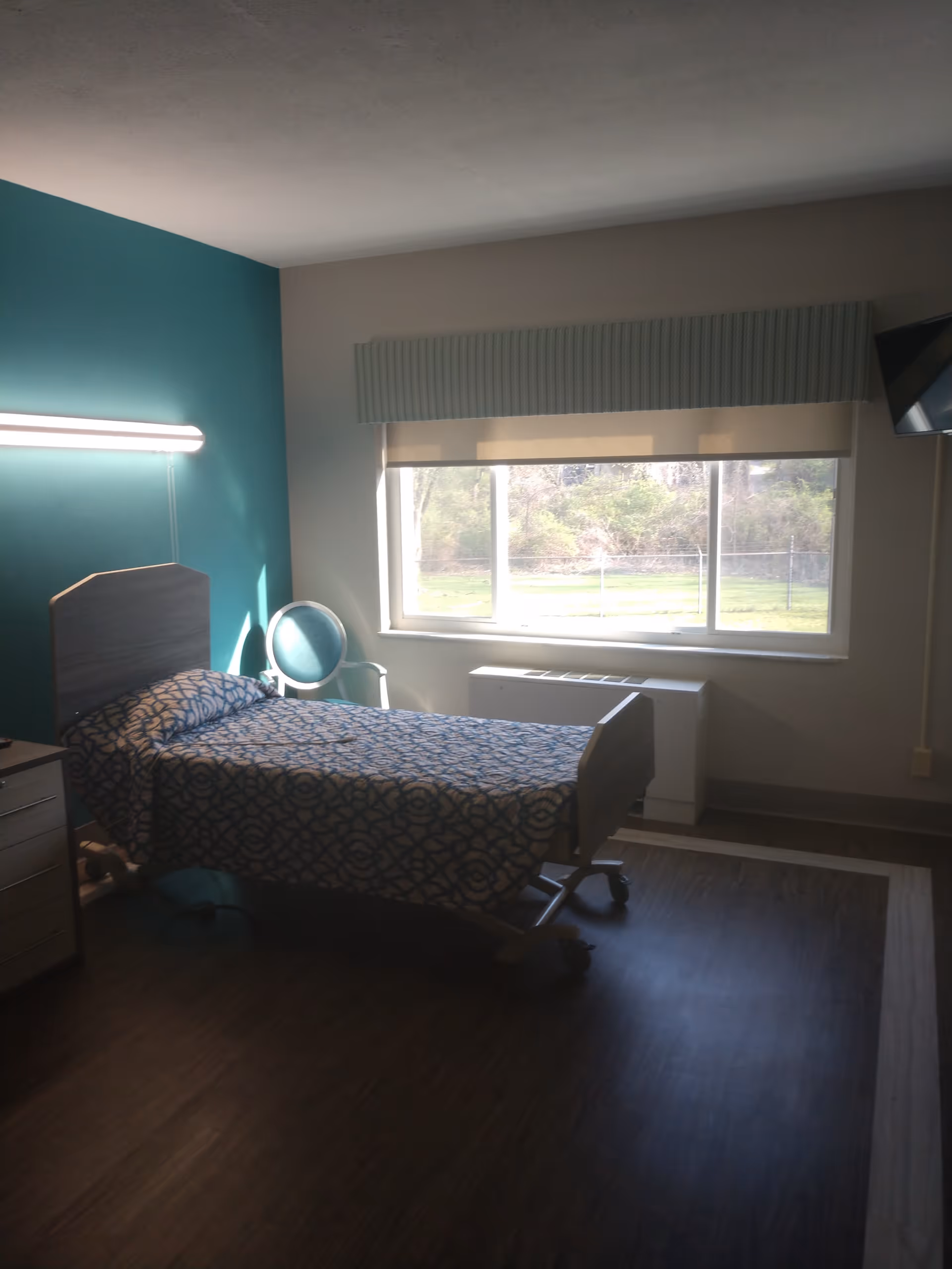 A simple, clean bedroom in a rehab and nursing facility with a single bed covered in a patterned bedspread, a teal accent wall, a round chair, a window with a view of greenery outside, and a wall-mounted TV in the corner.