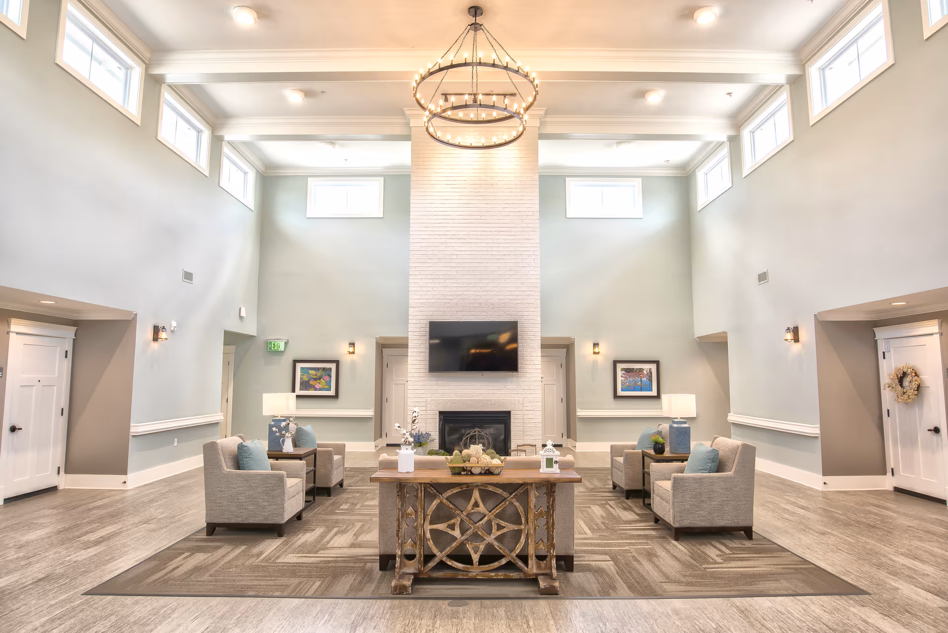 A spacious and well-lit senior living facility common area with high ceilings and multiple small windows near the ceiling. The room features a central white brick fireplace with a mounted flat-screen TV above it. There are four gray armchairs with light blue cushions arranged in pairs on either side of the fireplace, each pair accompanied by a side table with a lamp. A decorative wooden console table with ornamental details is placed behind a sofa facing the fireplace. The walls are painted light gray, and the floor has a combination of wood and carpeted areas. Two framed artworks hang on the wall behind the seating area, and there are white doors on either side of the fireplace.
