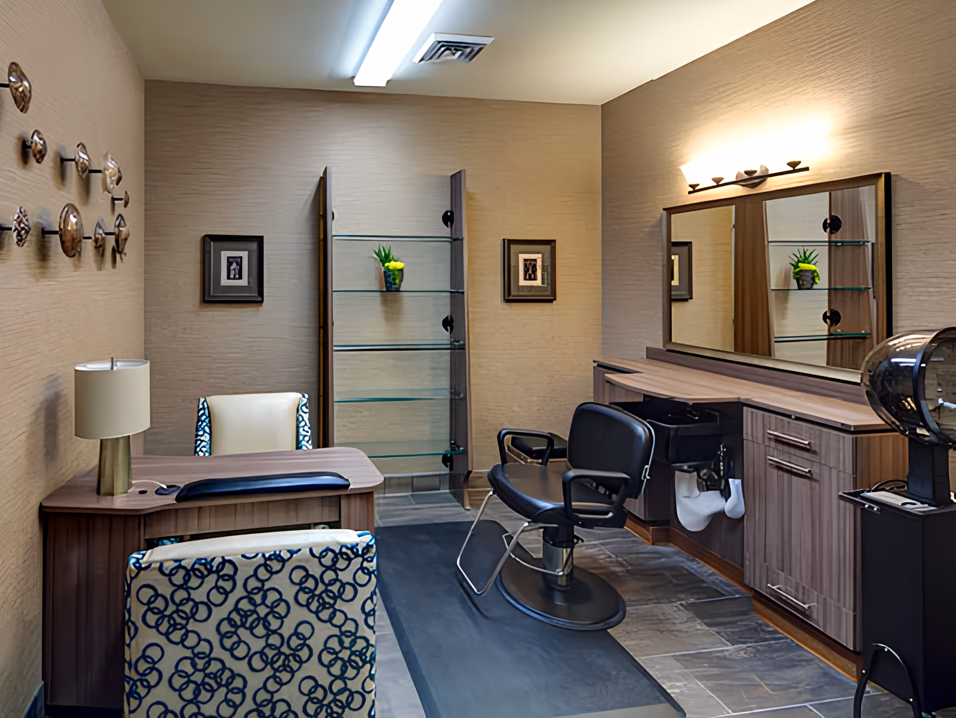 A well-lit salon room with a barber chair in front of a large mirror and wooden cabinetry. There is a desk with a lamp and two patterned chairs, along with decorative wall art and a glass shelving unit with a small plant.