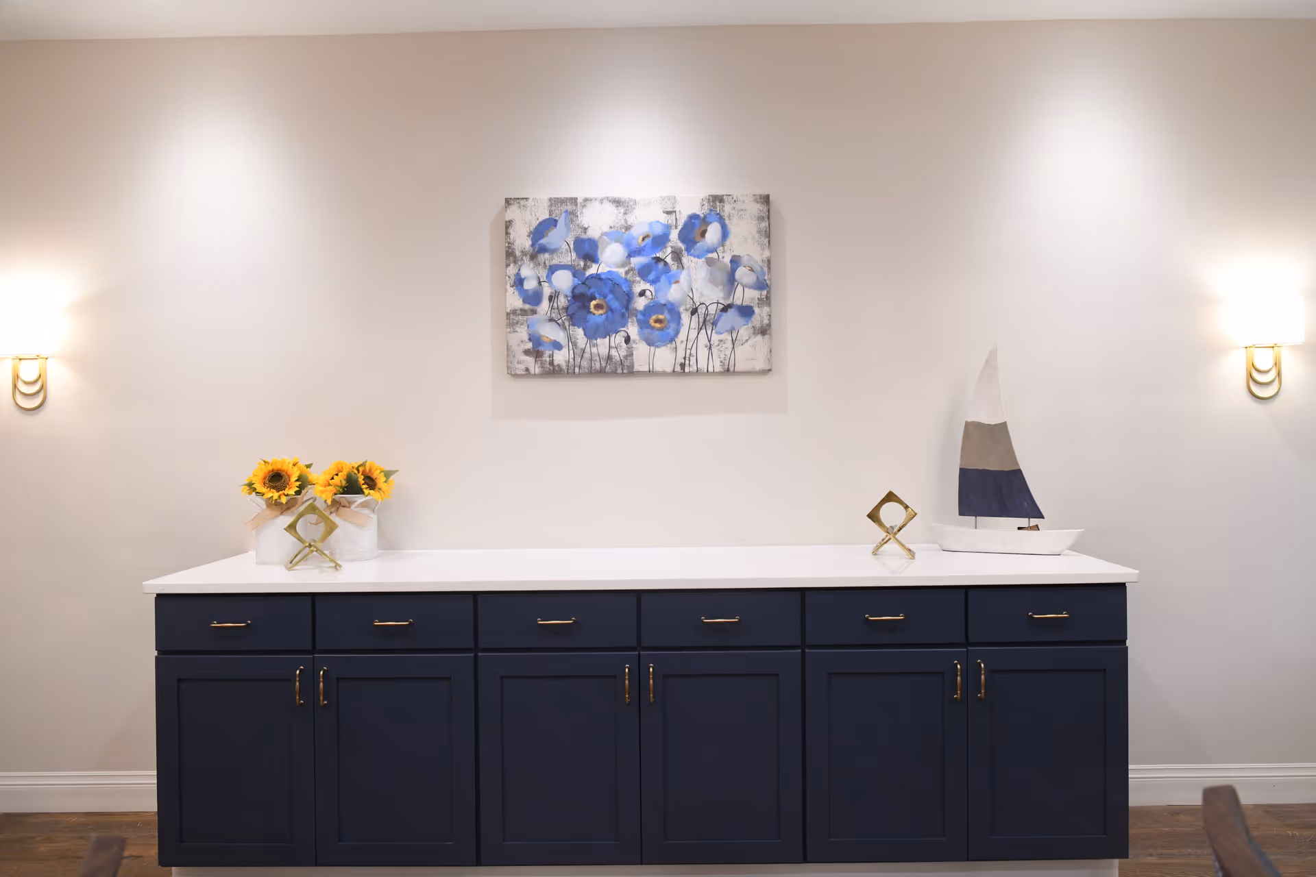 A navy blue cabinet with gold handles topped with a white countertop. On the left side of the countertop are two white vases with sunflowers and a small gold decorative object. On the right side is a white and blue sailboat decoration and another small gold decorative object. Above the cabinet is a painting of blue flowers on a white and gray background. The wall is light-colored with two wall sconces emitting warm light on either side of the cabinet.