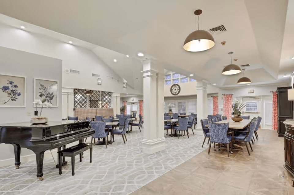 A spacious, well-lit common area with multiple tables and blue upholstered chairs arranged for dining or socializing. The room features a black grand piano on the left, decorative wall art, large windows with red patterned curtains, and pendant lights hanging from the ceiling. White columns and a large clock on the far wall add to the elegant atmosphere.