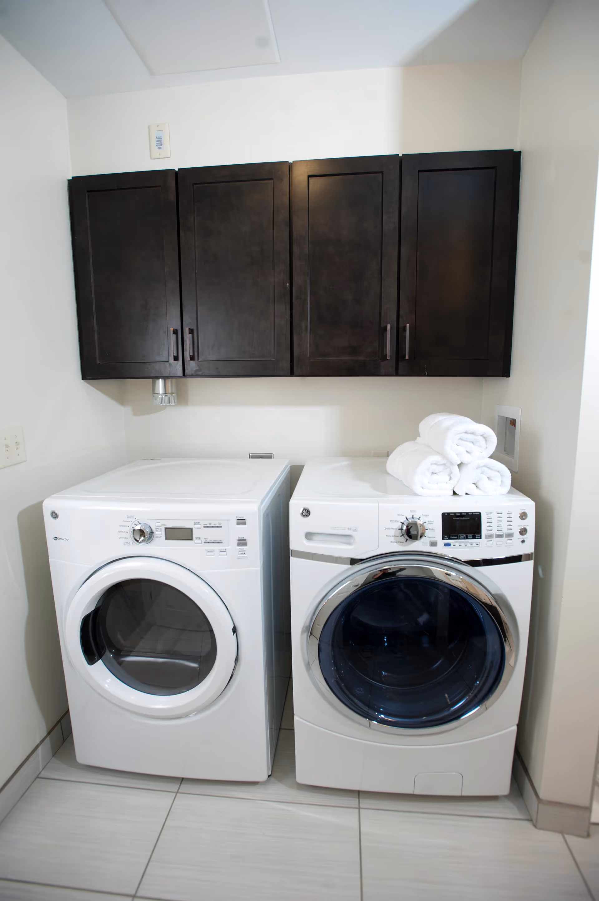 A laundry room with a white front-loading washing machine and dryer side by side. Above them are dark wooden cabinets mounted on the wall. Three neatly rolled white towels are placed on top of the dryer. The room has light-colored tiled flooring and white walls.