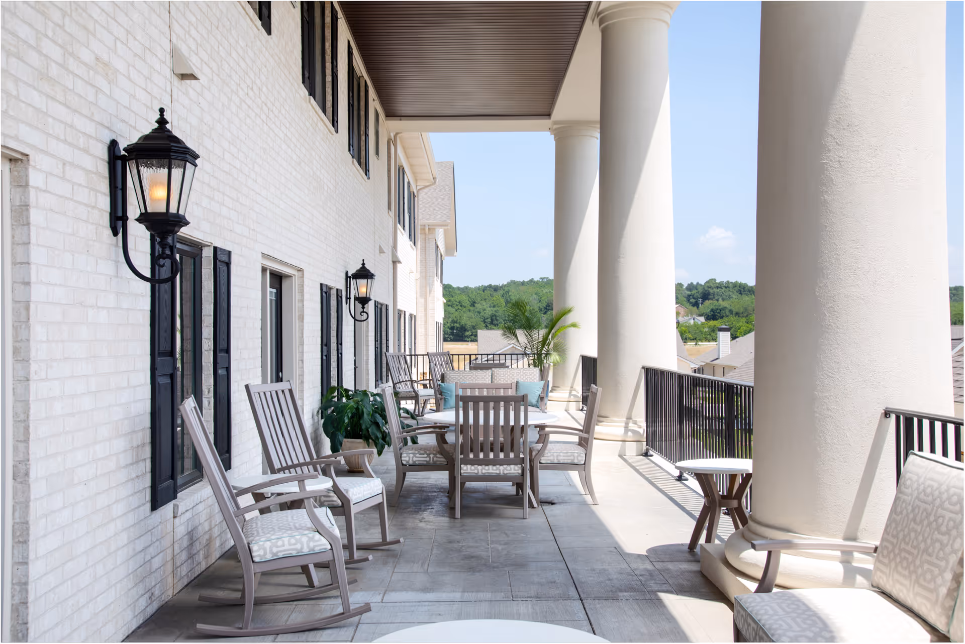 A spacious outdoor balcony area with large white columns, several cushioned chairs, a round table, potted plants, and black wall-mounted lanterns on a white brick wall. The balcony overlooks a scenic view of trees and rooftops under a clear blue sky.