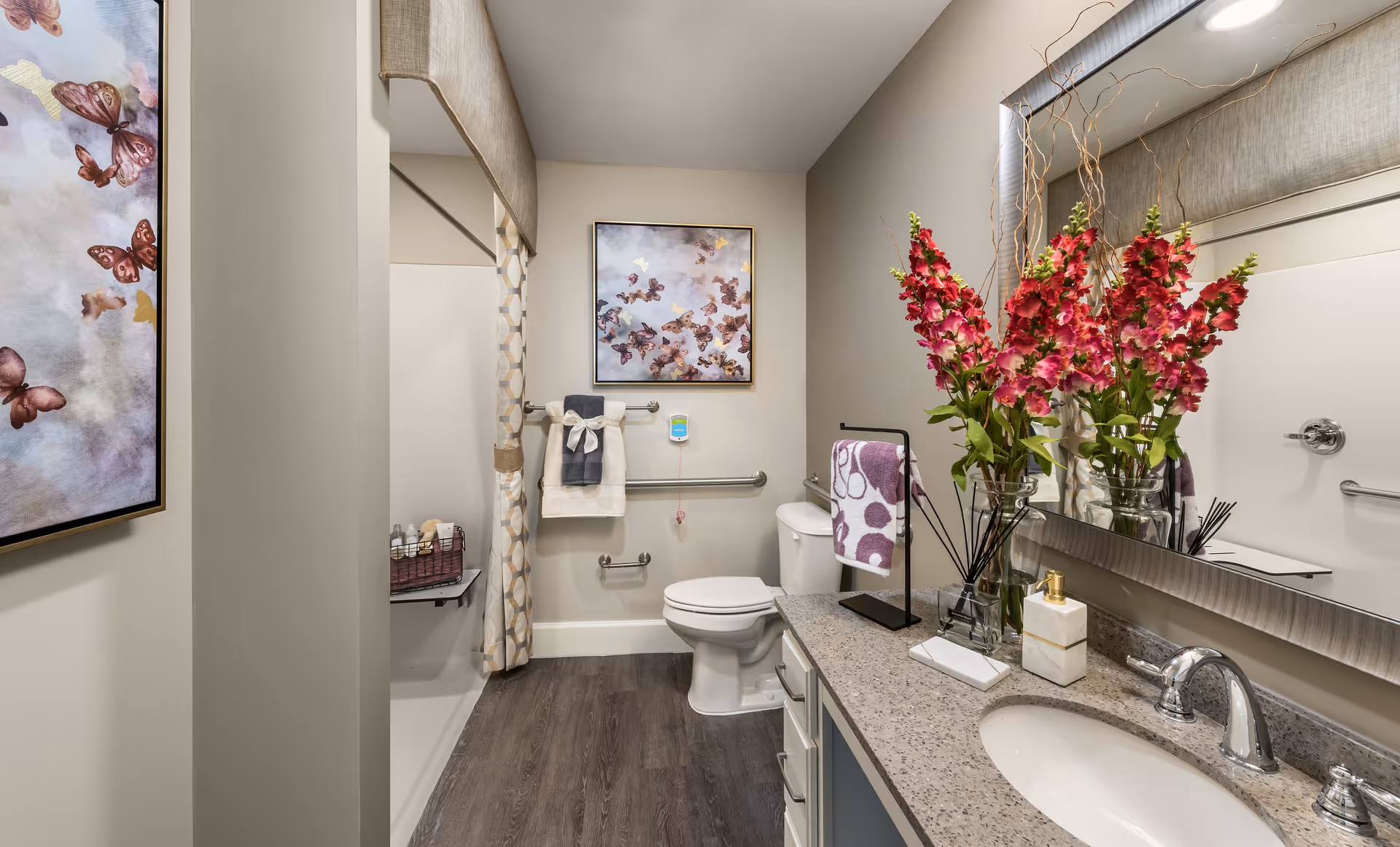 A clean and well-decorated bathroom featuring a toilet, a sink with a granite countertop, a large mirror, and a shower with a patterned curtain. The countertop has a vase with vibrant pink flowers, a soap dispenser, and a small towel rack with a purple and white towel. The walls are adorned with a framed butterfly artwork and there are grab bars installed near the toilet and shower for accessibility.
