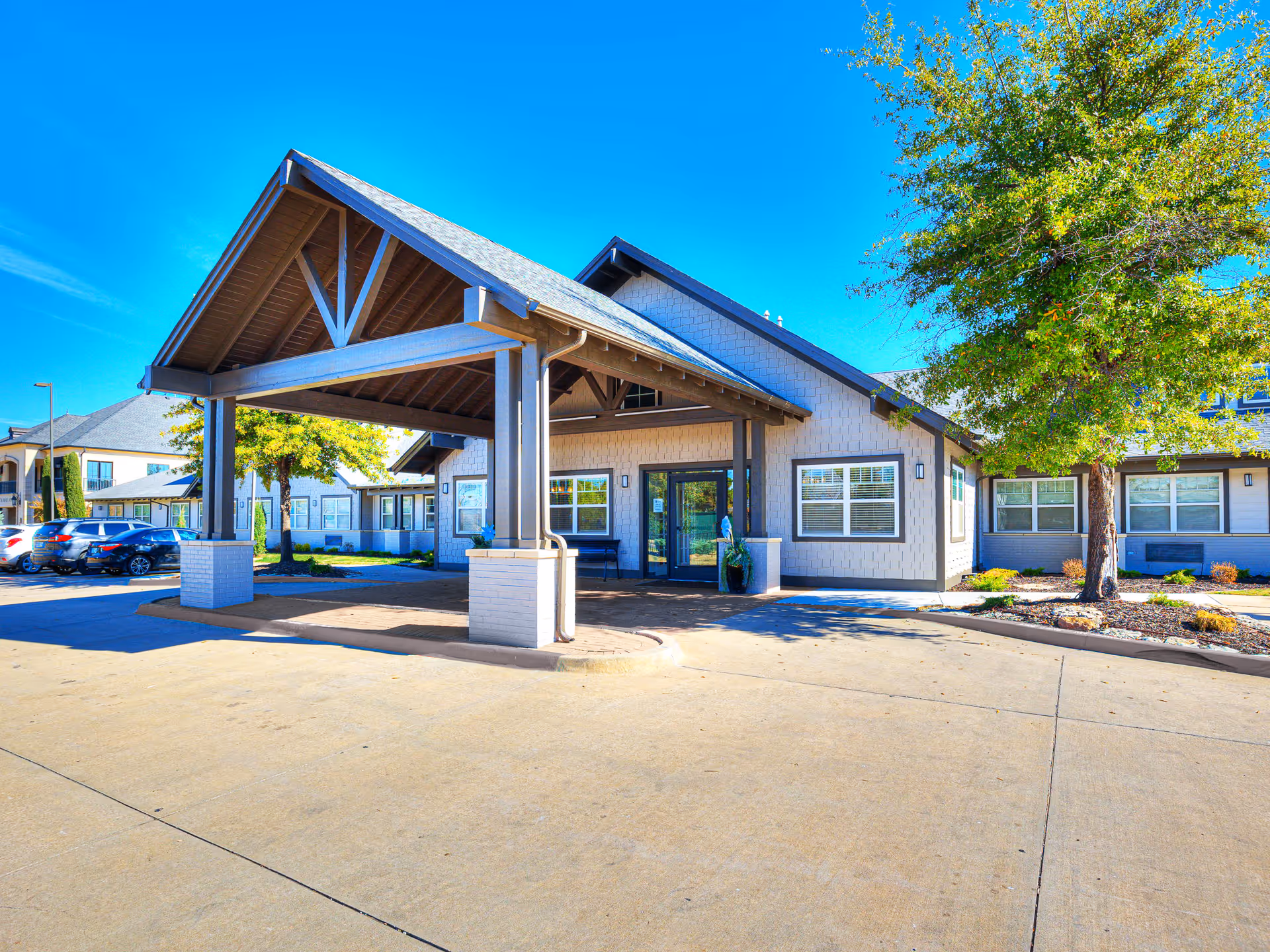 Front entrance of a single-story senior living building with a large covered porte-cochere and a tree to the right.