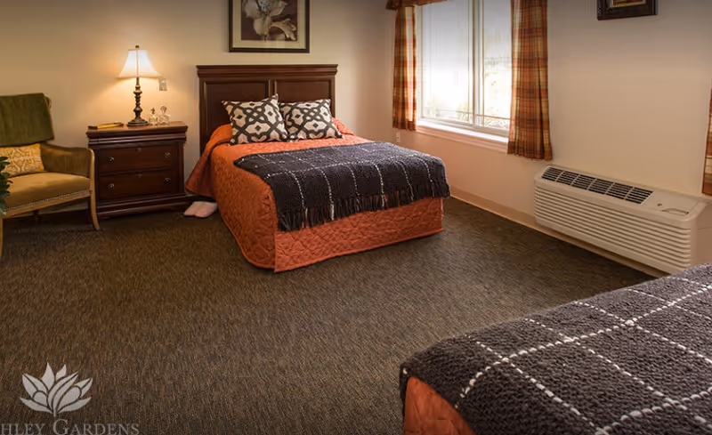 A cozy bedroom in an assisted living facility featuring two beds with orange bedspreads and dark knitted throws. There is a wooden nightstand with a lamp and decorative items between the beds, a comfortable armchair with a green throw, a large window with plaid curtains, and a wall-mounted air conditioning unit below the window.