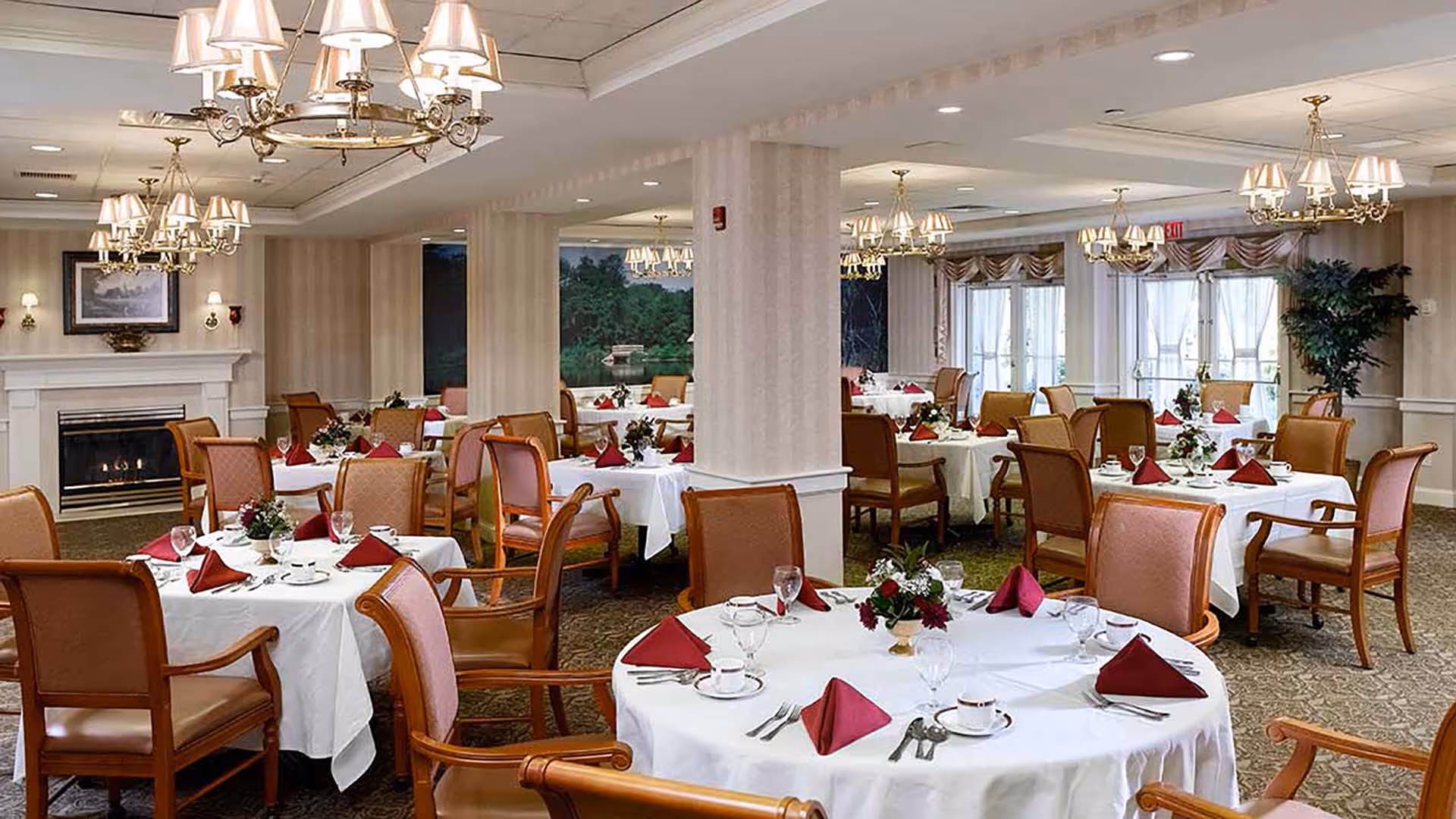 A spacious dining room with multiple round tables covered in white tablecloths, each set with red folded napkins, glassware, and floral centerpieces. The room features elegant chandeliers, a fireplace with a framed picture above it, large windows with curtains, and comfortable wooden chairs with padded seats and backs.