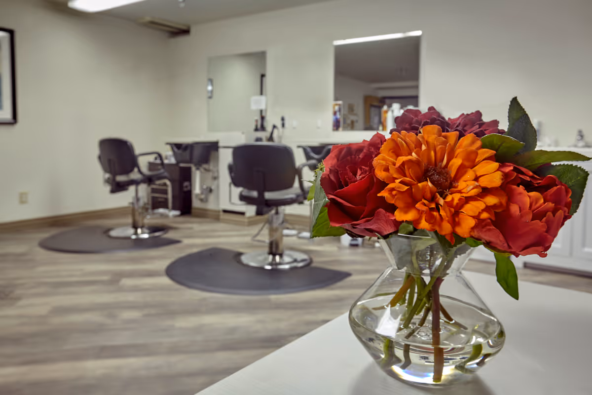 A bright and clean salon area with two black salon chairs in front of large mirrors mounted on the wall. In the foreground, there is a clear glass vase with a bouquet of vibrant orange and red flowers placed on a white surface. The room has light-colored wooden flooring and white walls.