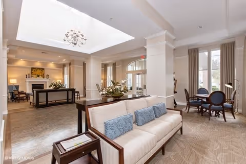 A spacious and well-lit living room area in a senior living facility featuring a beige sofa with blue cushions, a wooden side table with magazines, a chandelier hanging from a recessed ceiling, a fireplace with decorative items, and a round table with chairs near large windows with curtains.