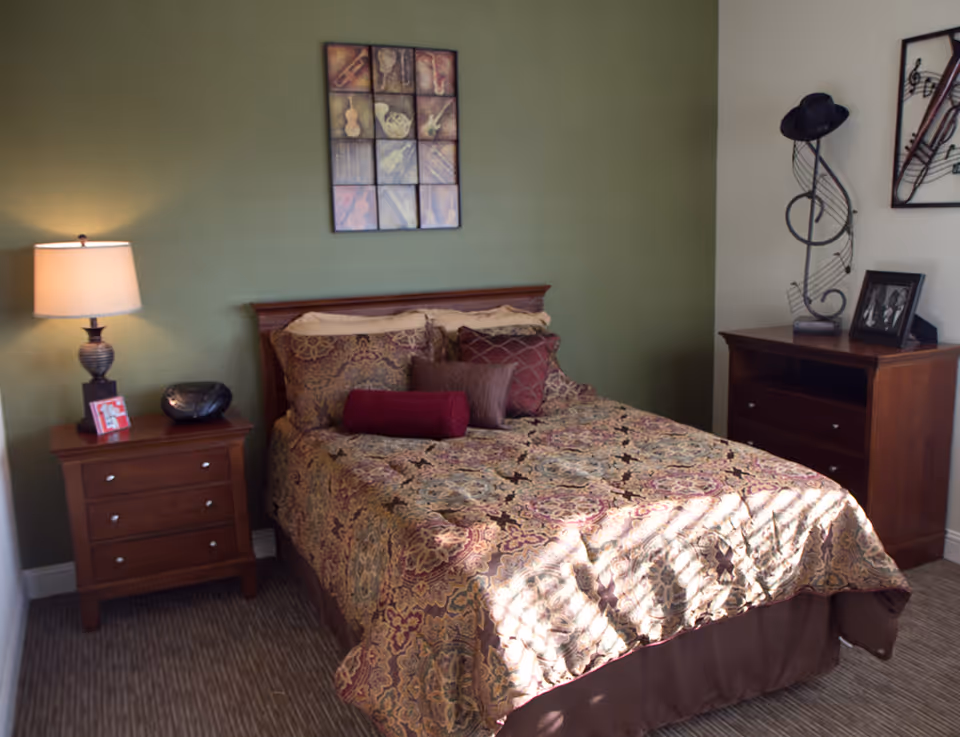 A cozy bedroom with a double bed covered in a patterned bedspread and multiple pillows. There is a wooden nightstand with a lamp, a small radio, and a framed photo on it. On the wall above the bed is a framed artwork featuring musical instruments. To the right of the bed is a wooden dresser with a decorative metal music note sculpture and a framed photo. The walls are painted green and white, and sunlight is streaming onto the bed.