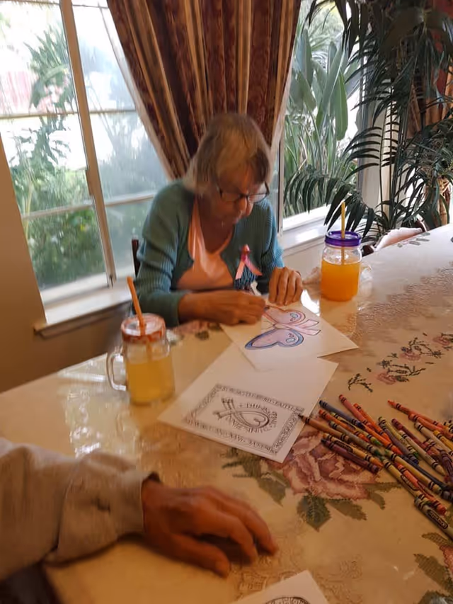 An elderly woman wearing glasses and a teal cardigan is sitting at a table by a window, coloring a butterfly drawing with crayons. There are two drinks with straws on the table, along with several crayons and other coloring sheets. The table has a floral tablecloth and there are plants visible near the window.