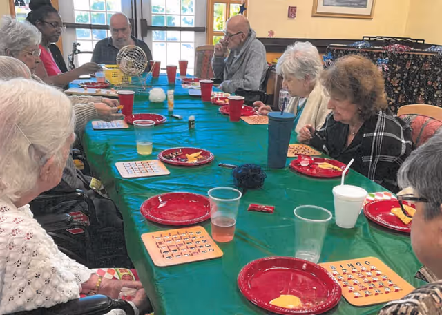 A group of elderly people sitting around a long table covered with a green tablecloth, playing bingo. There are red plates with snacks, plastic cups with drinks, and bingo cards on the table. The room has large windows letting in natural light.