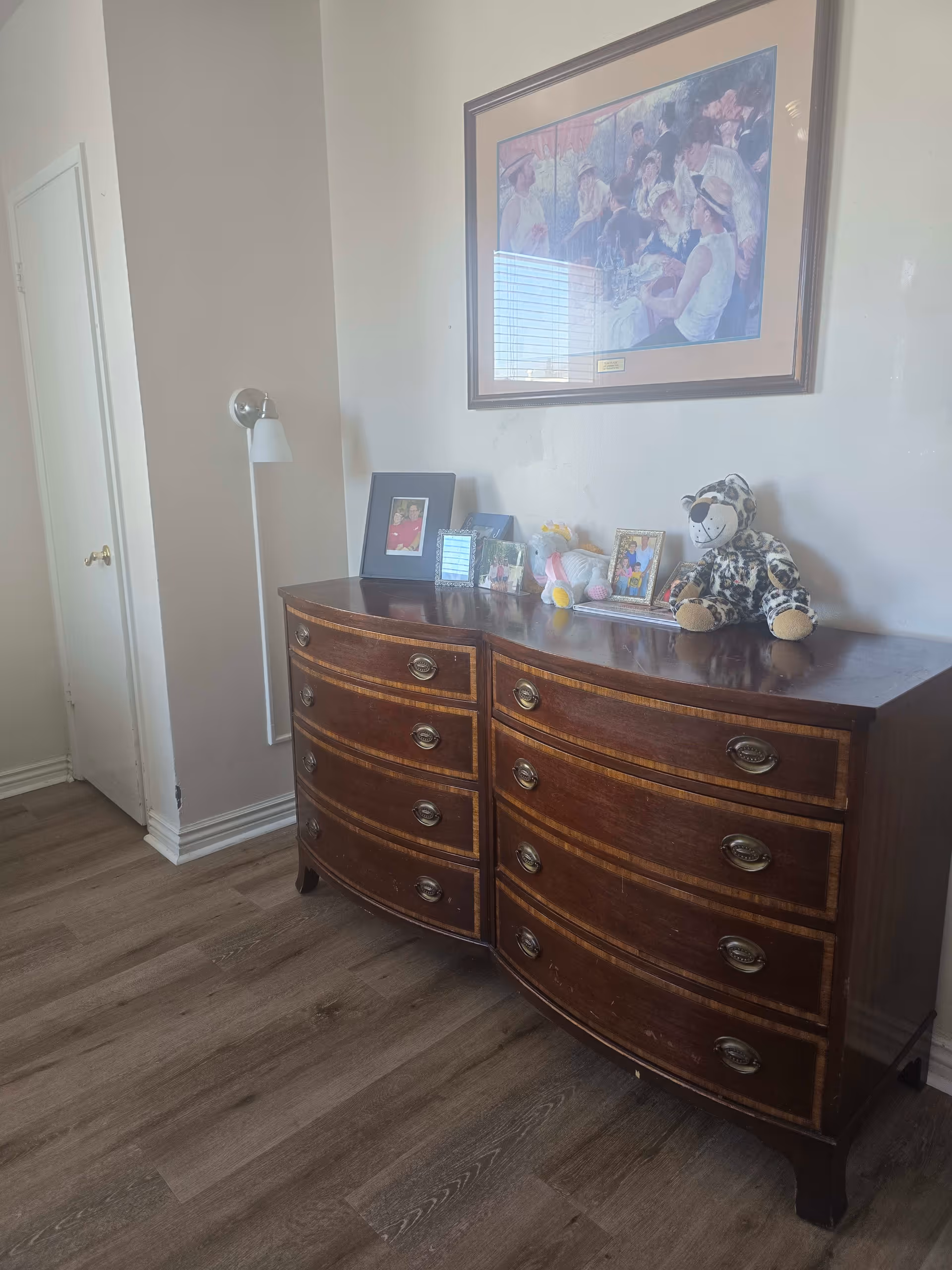 A wooden dresser with curved front and multiple drawers, topped with framed photos, a stuffed animal, and a plush toy. Above the dresser hangs a framed painting depicting a group of people gathered outdoors. The room has light-colored walls, a wooden floor, a closed white door, and a wall-mounted lamp beside the dresser.