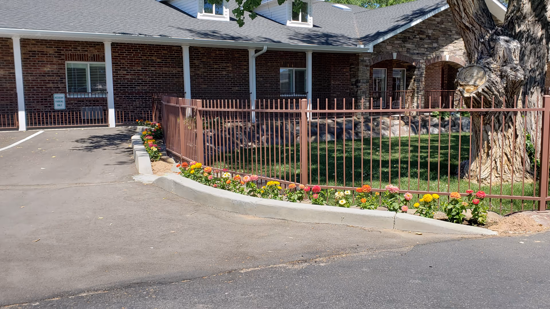 Exterior view of BeeHive Homes of Bosque Farms showing a brick and stone building with a fenced grassy area and colorful flowers planted along the fence. There is a large tree on the right side and a parking area on the left with a sign indicating reserved parking.