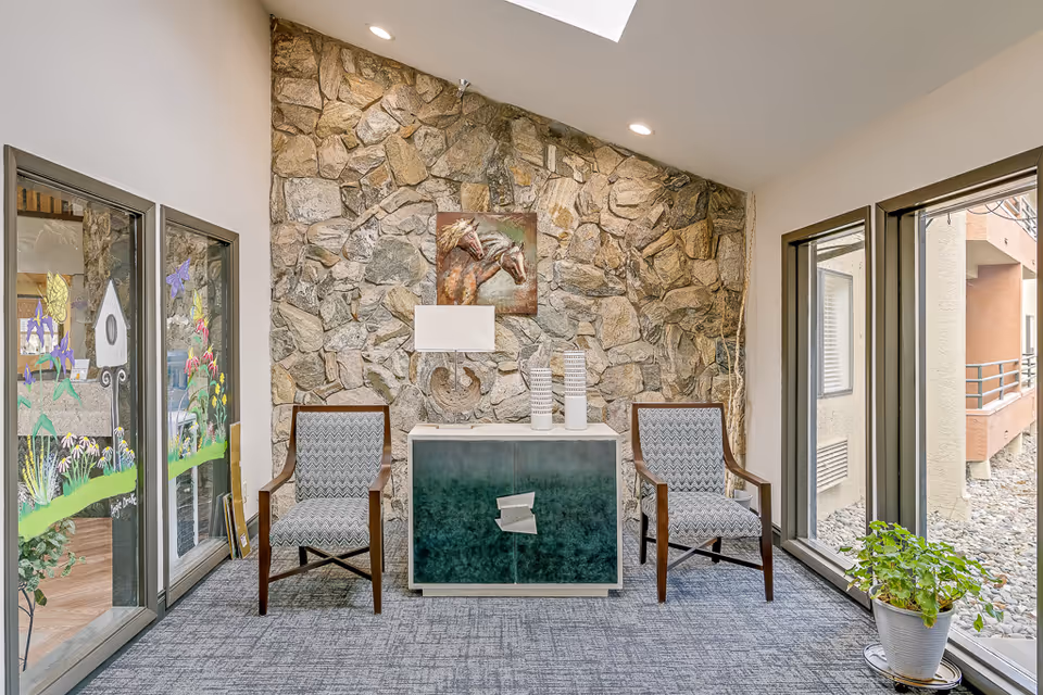 Small seating area with two upholstered chairs flanking a green cabinet against a stone accent wall, large windows and a potted plant.