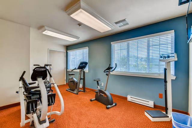 A small exercise room with orange carpet and light blue walls, containing stationary exercise bikes, a rowing machine, and a standing scale. Two large windows with white blinds allow natural light into the room.