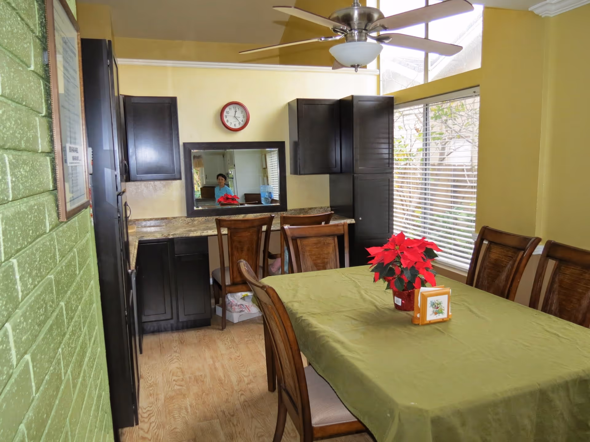 Dining room with a rectangular table covered with a green tablecloth and surrounded by wooden chairs. A red poinsettia plant and a small framed picture are on the table. The room has large windows with blinds, yellow walls, and dark wooden cabinets along one wall. A ceiling fan with a light fixture is mounted above the table. A clock and a mirror are on the wall above a countertop with chairs underneath. A person is partially visible in the mirror reflection.