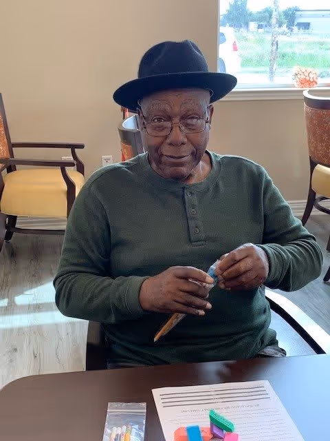 An elderly man wearing a dark hat and green long-sleeve shirt is sitting at a table in a well-lit room with wooden flooring and chairs. He is holding a pencil and some small objects in his hands. On the table in front of him are colorful blocks and a sheet of paper with text. A window in the background shows a view of the outdoors.