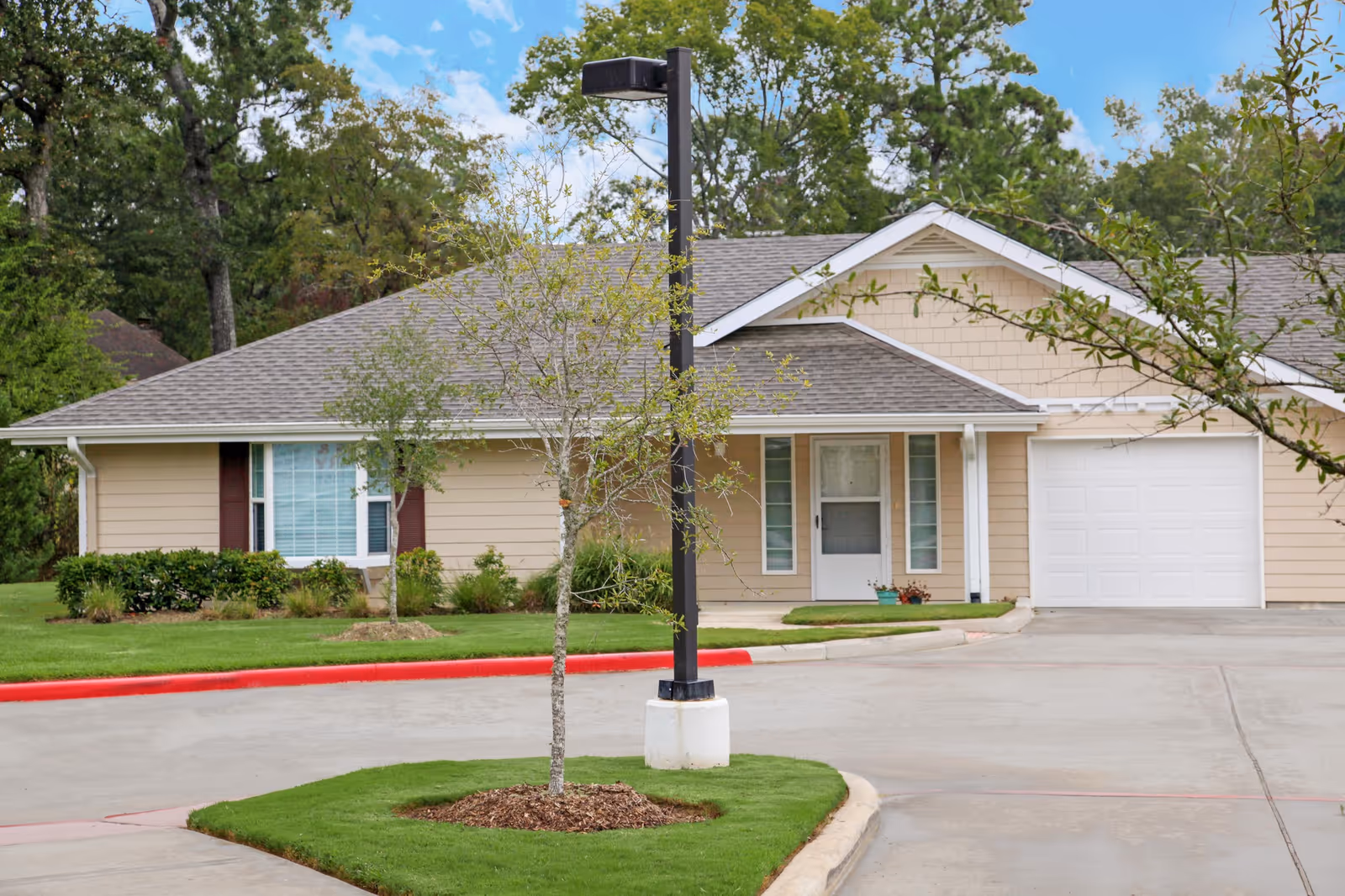Single-story beige residential building with a white garage door and a front entrance door, surrounded by green grass, small trees, and bushes under a partly cloudy sky.
