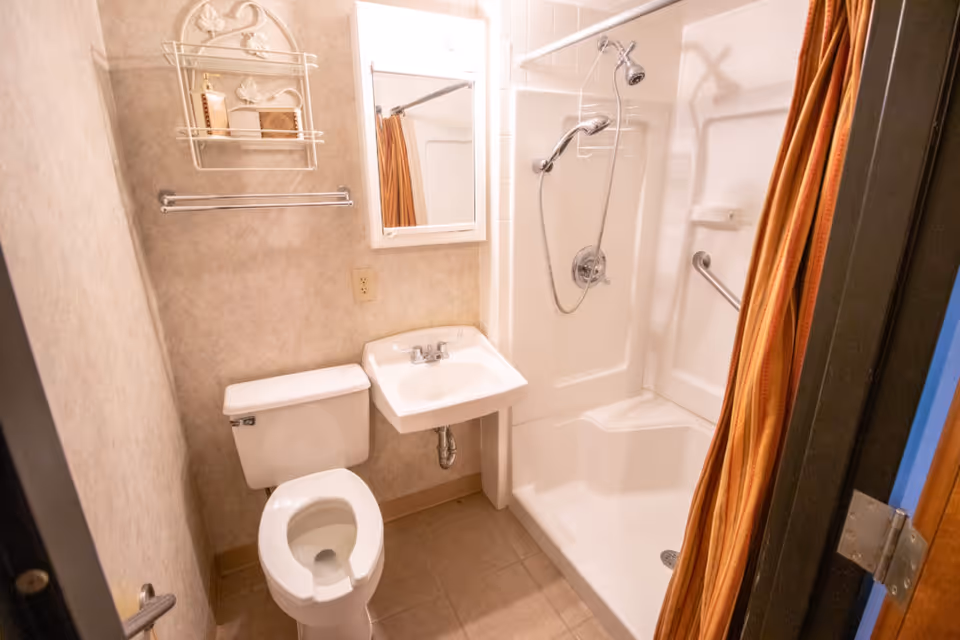 Small bathroom with a white toilet, a white sink with a mirror above it, and a shower with a handheld showerhead and a built-in seat. There is a towel rack on the wall and a decorative shelf holding toiletries. The shower curtain is orange with stripes.