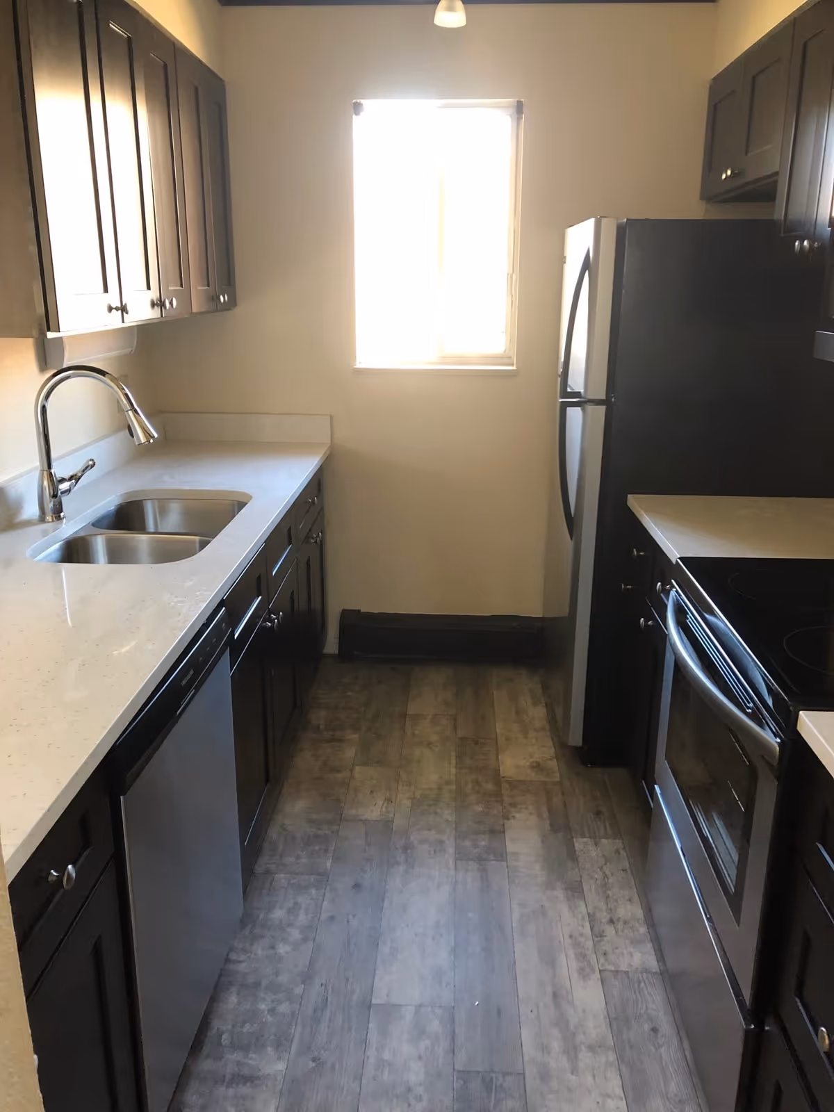 A narrow kitchen with dark wood cabinets, a light-colored countertop, a stainless steel sink with a modern faucet, a dishwasher, a stainless steel refrigerator, and a stainless steel electric stove. There is a window at the end of the kitchen letting in natural light, and the floor has a wood-like finish.