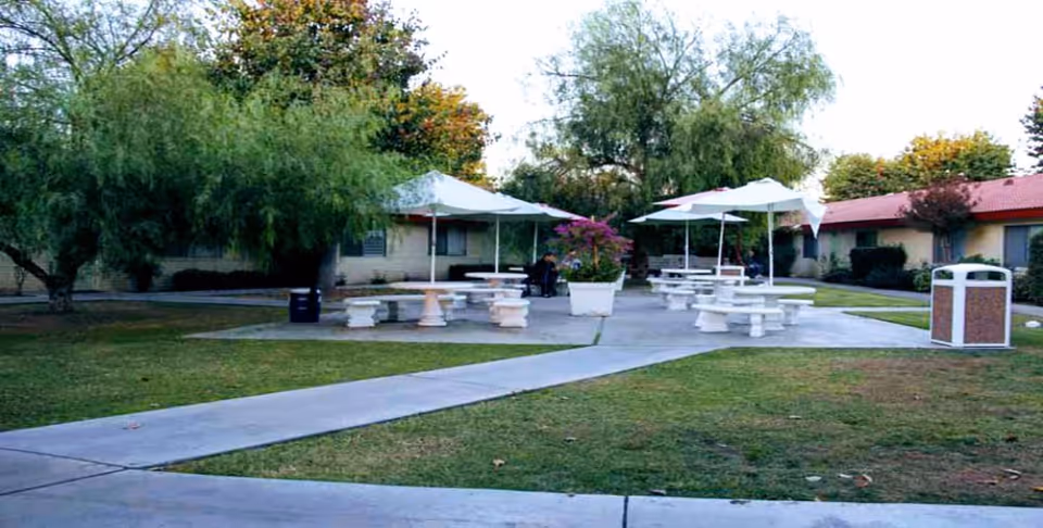 Outdoor courtyard with picnic tables and umbrellas on paved pads, surrounded by grass, trees, and single-story buildings.