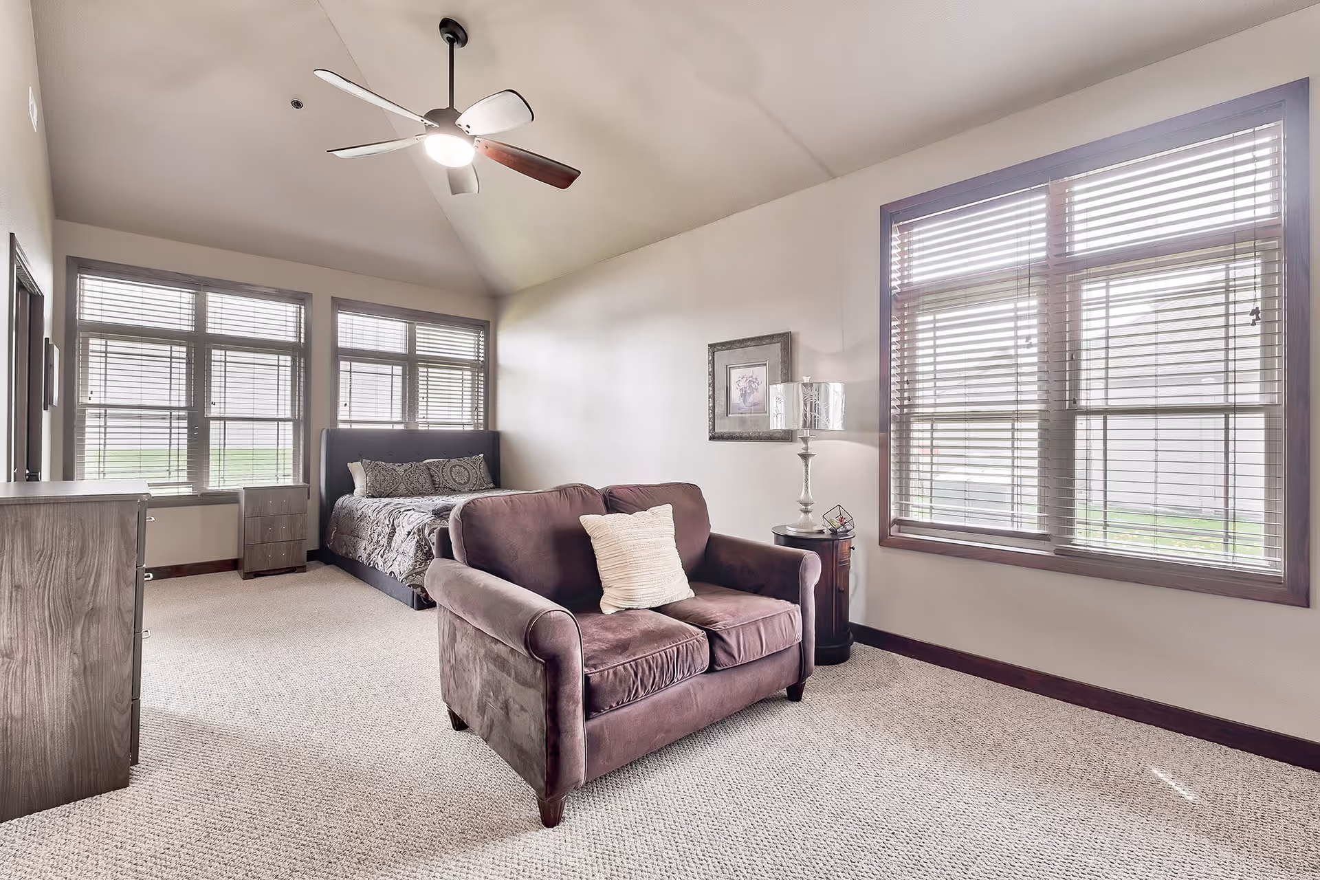 A bright and spacious bedroom with a large window featuring wooden blinds. The room includes a bed with patterned bedding, a small loveseat with a white pillow, a wooden dresser, a nightstand with a lamp, and a ceiling fan with a light fixture. The walls are light-colored, and the carpet is beige.