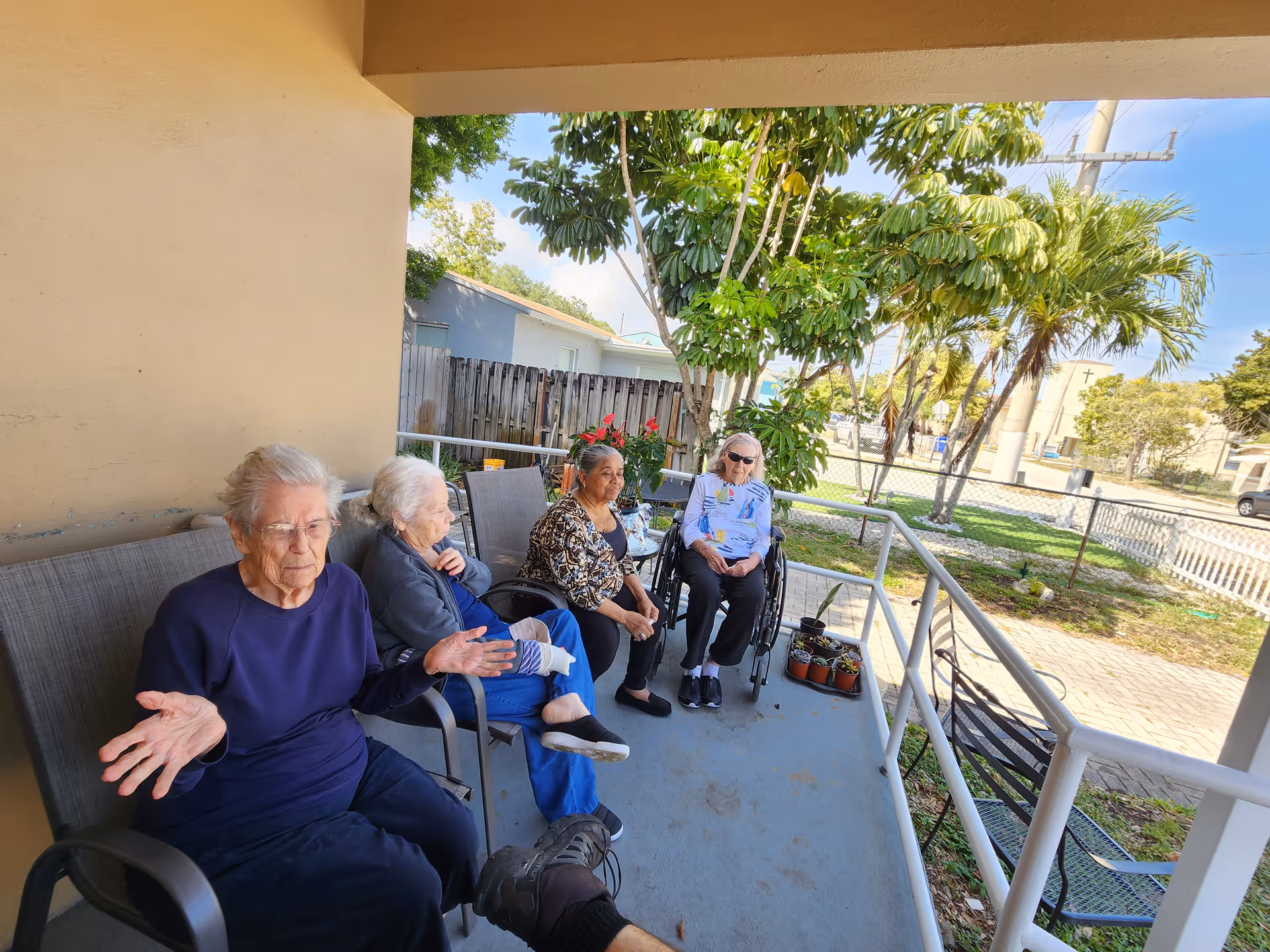 Four elderly women sitting on chairs on a covered outdoor patio. One woman is in a wheelchair. There are potted plants on the ground and trees and a fence in the background under a clear blue sky.