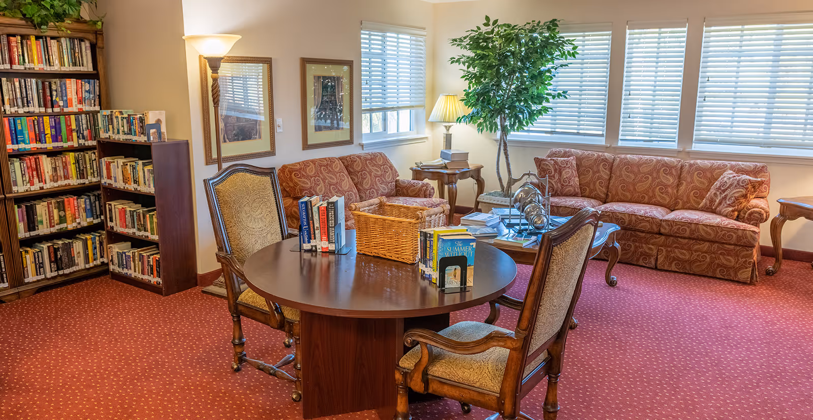 A cozy living room area with two patterned sofas, a round wooden table with chairs, and a bookshelf filled with books. The room has red carpet flooring, several windows with blinds, a floor lamp, a table lamp, framed pictures on the wall, and a large potted plant near the windows.