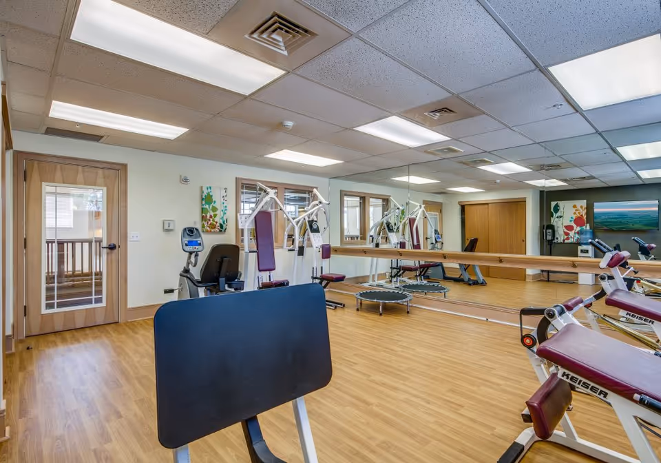 Bright exercise room with workout machines, a mirrored wall, wooden floor, and ceiling lights.