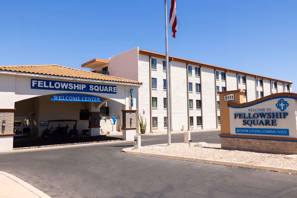 Exterior view of Fellowship Square Tucson senior living community showing the entrance with a covered driveway labeled 'Fellowship Square Welcome Center' and a large sign that reads 'Welcome to Fellowship Square Senior Living Community' with the address 8111. The building is a multi-story structure with multiple windows and a clear blue sky above.