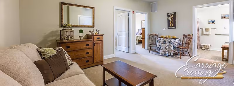A cozy living room area with a beige sofa adorned with patterned cushions, a wooden coffee table, and a wooden dresser with decorative items and a large mirror above it. In the background, there is a small dining area with a round table covered with a floral tablecloth and two wooden chairs. Two open doorways lead to a bedroom and a bathroom, both visible in the image. The walls are painted light beige, and the carpet is light-colored.