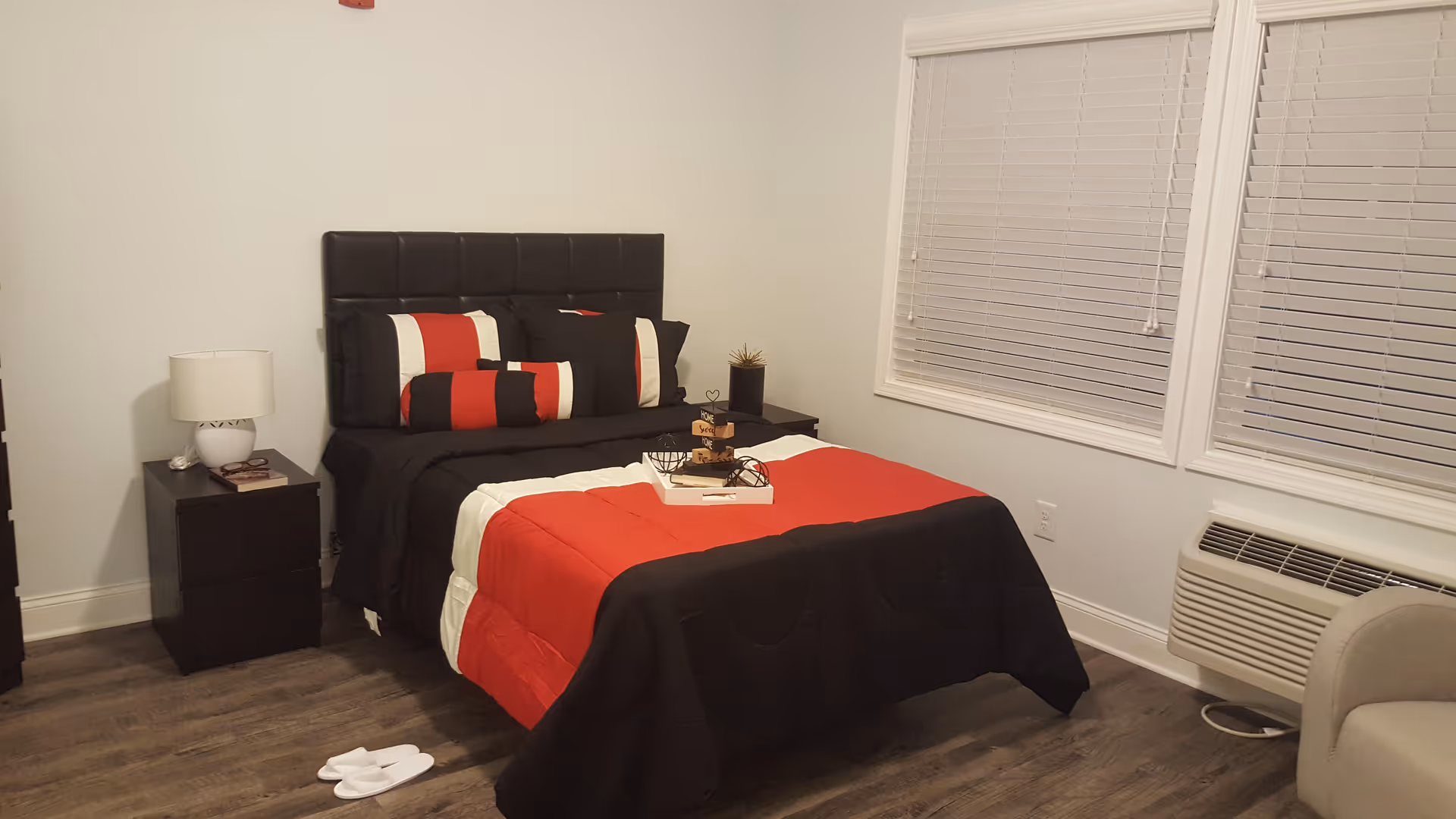 A neatly made bed with a black, red, and white comforter and matching pillows in a bedroom. The room has wooden flooring, two nightstands with a lamp and decorative items, two windows with closed blinds, a wall-mounted air conditioning unit, and a beige armchair partially visible on the right.