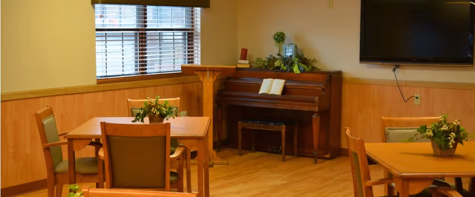 Small communal room with wooden tables and chairs, a piano against the wall, a wall-mounted TV, and potted plants.