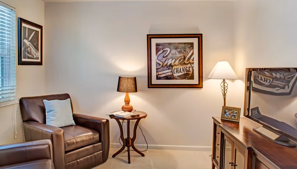 A cozy living room corner with two brown leather armchairs, a small wooden side table with a lamp and an open book, a framed artwork on the wall, a floor lamp, a wooden TV stand with a flat-screen TV, and a small framed photo.